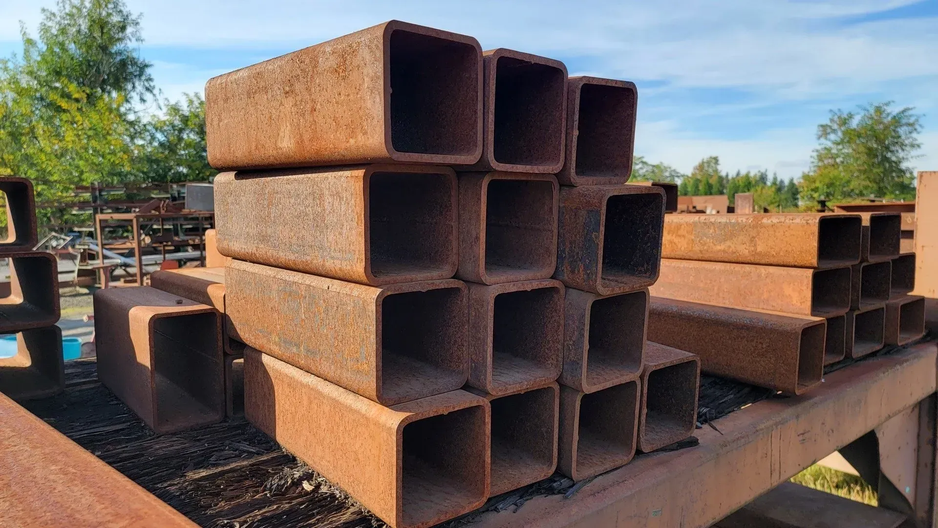 Rusty square steel tubes stacked outdoors under a blue sky.
