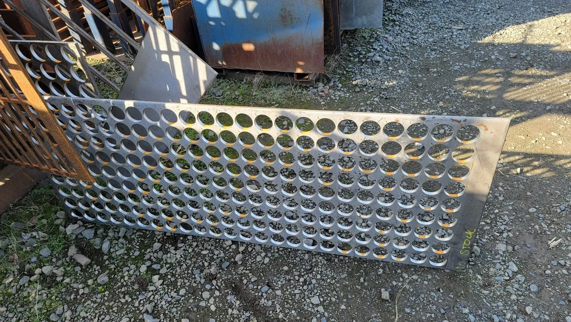 Metal sheet with evenly spaced circular holes. It rests on a gravel surface with other metal objects nearby.