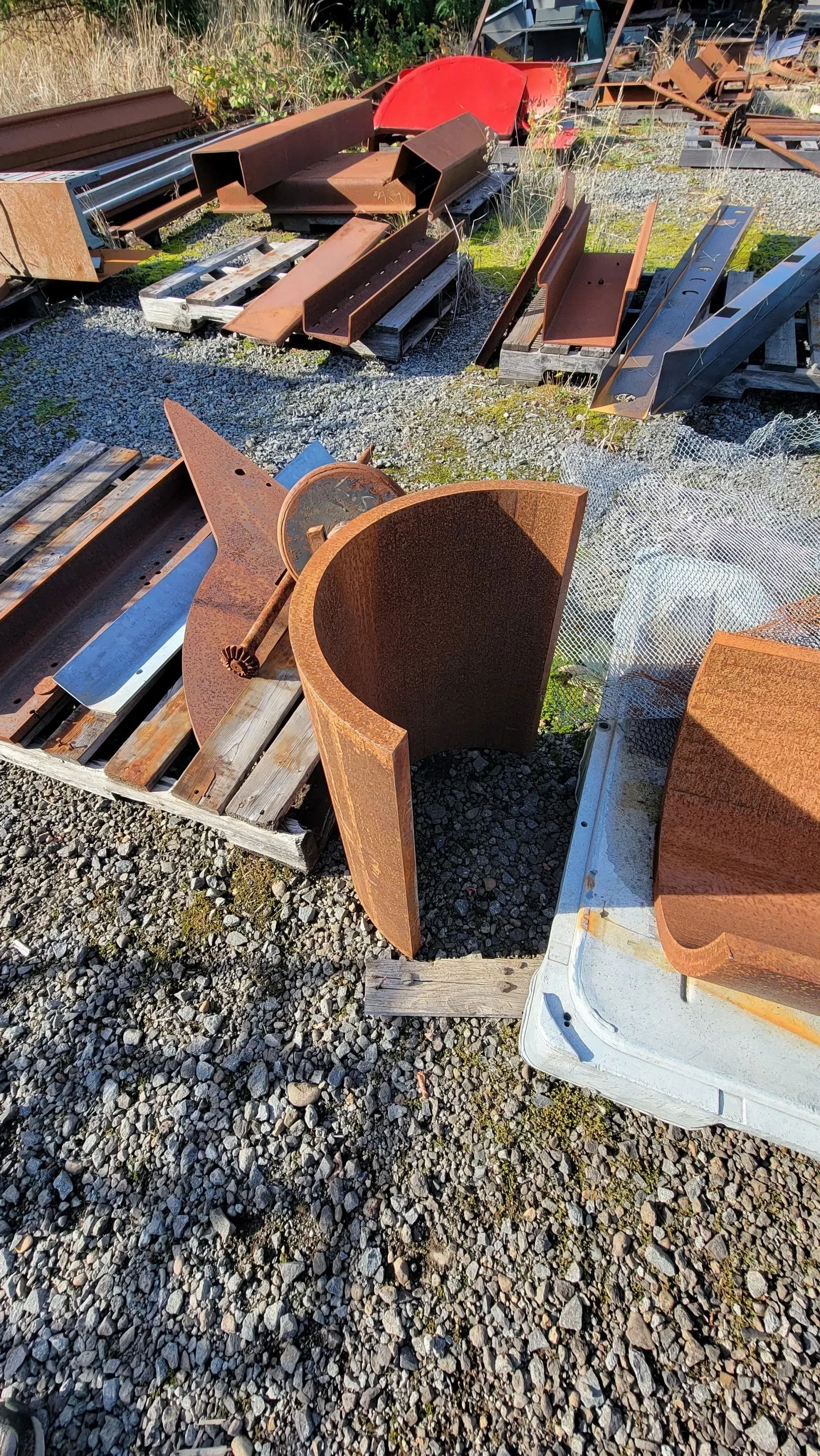 Rusty metal scraps and building materials on gravel. A curved, cylindrical piece of metal is prominent.