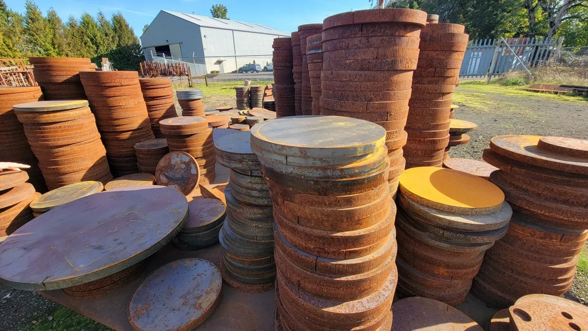 Stacks of rusty, round metal discs of varying sizes are outdoors with a building in the background.