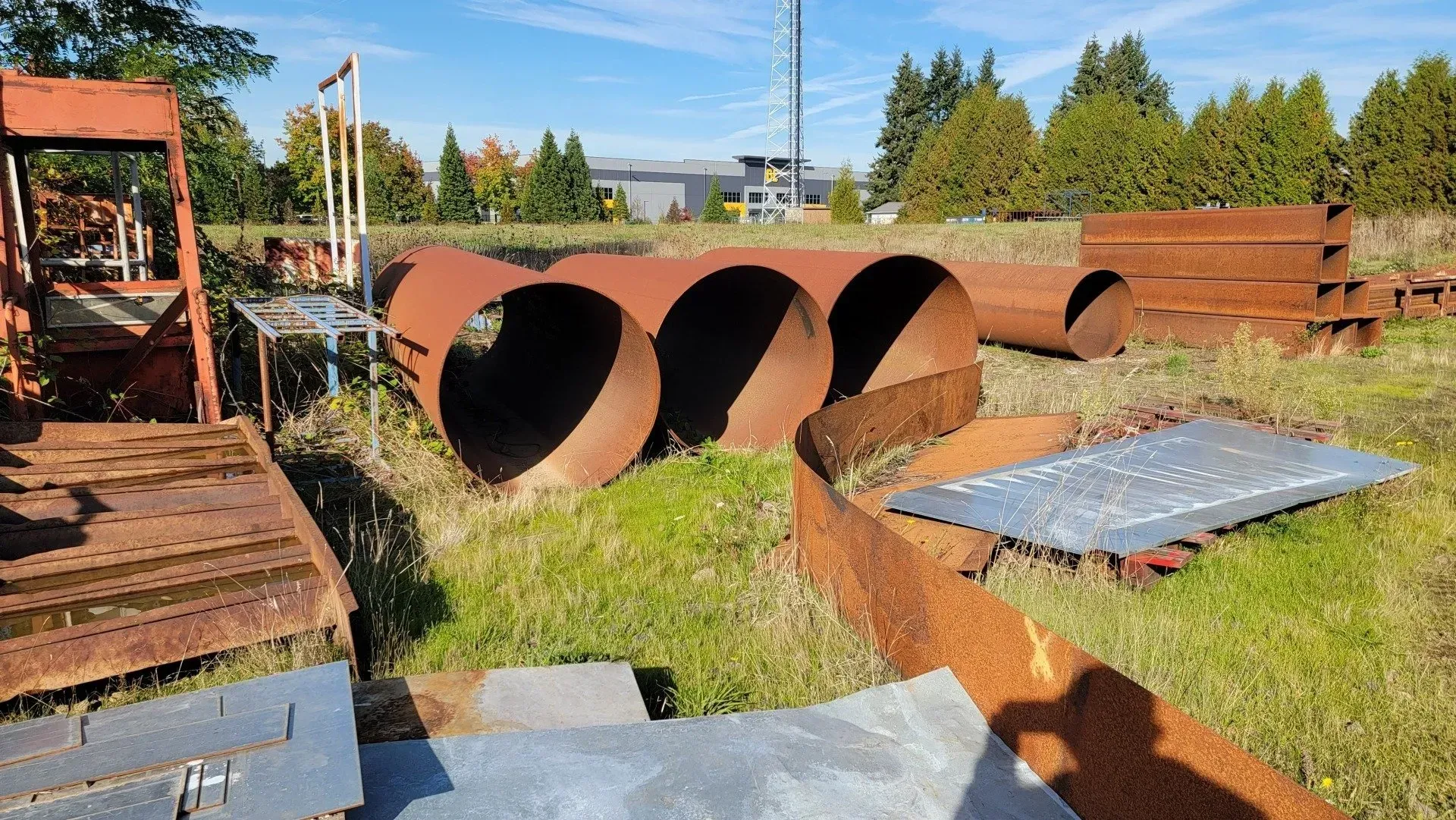 Rusty metal pipes and sheets of metal sit in a grassy field on a sunny day. Background includes trees and buildings.