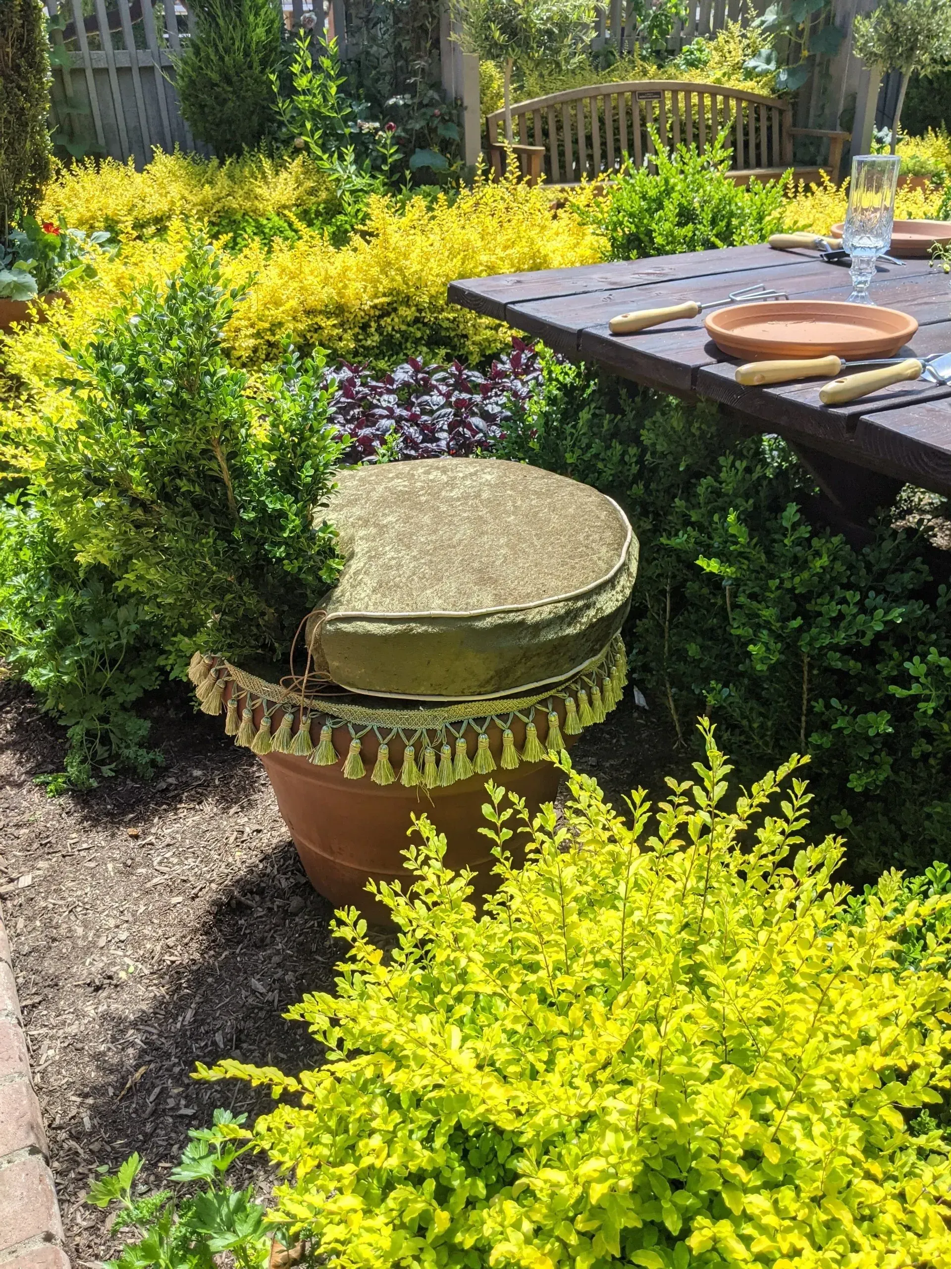 A terracotta pot with a gold cushioned seat in a garden setting. A dark wood table with tableware is in the background.