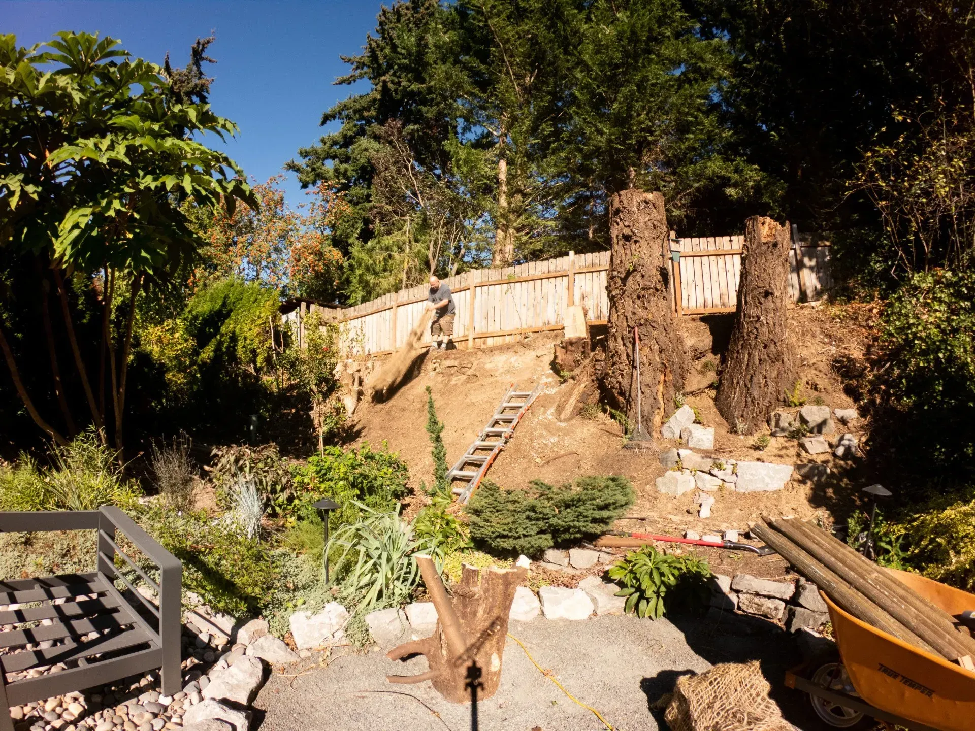 A backyard scene with a wooden fence atop a hillside, trees, and a seating area in the foreground.