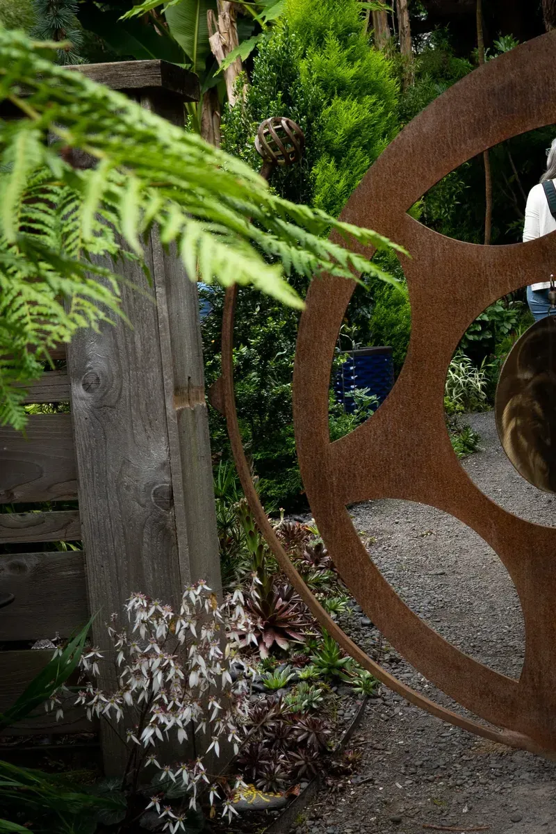 Rusty metal wheel gate in a garden, next to a wooden fence and greenery. A person is walking on a gravel path.