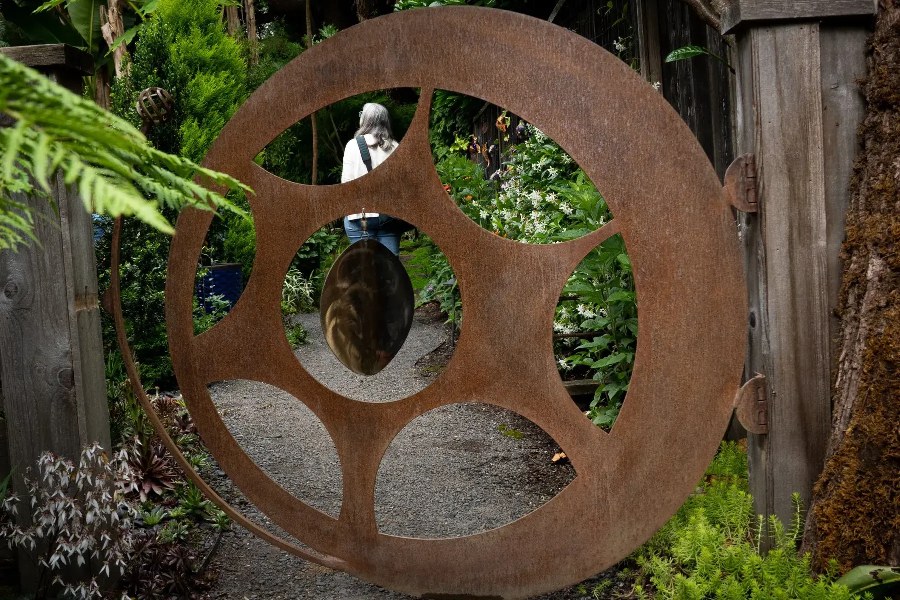 Rusty metal sculpture with cut-out design, mounted on a wooden fence with a blurred garden view in the background.