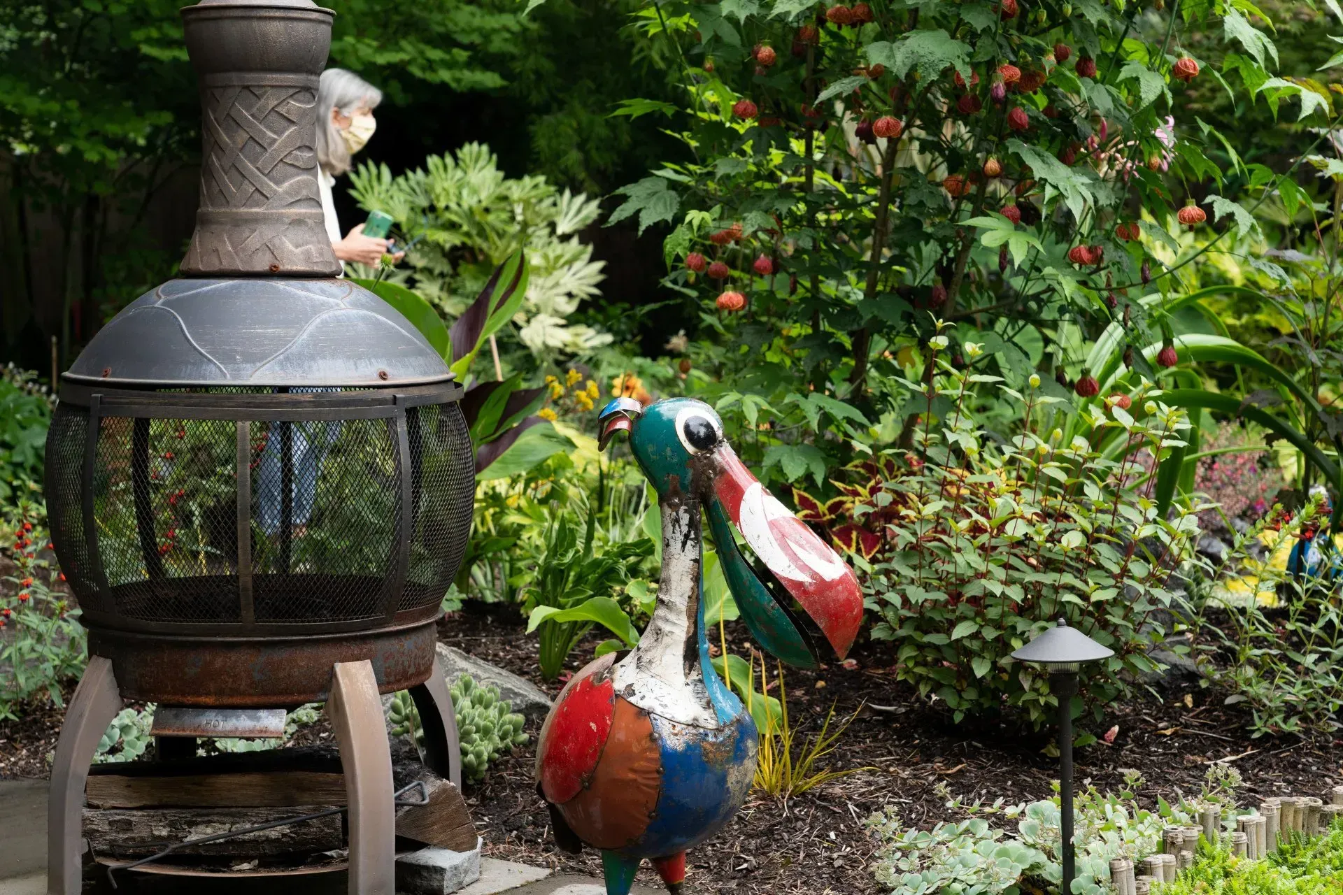 A metal bird statue and fire pit in a lush garden, person in the background.