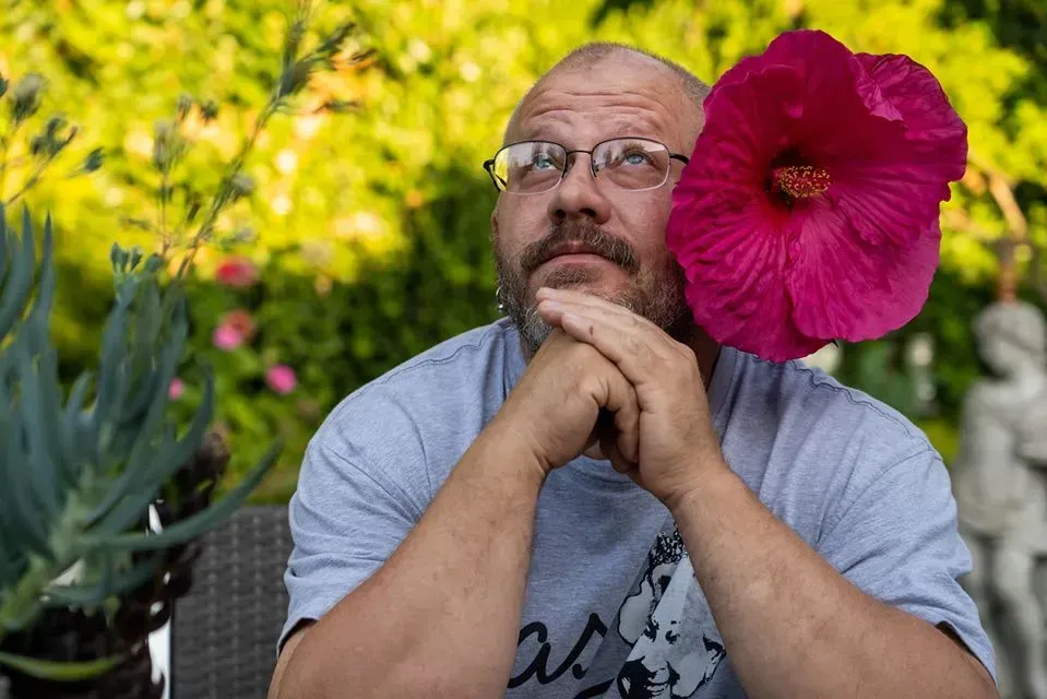 Man with a beard wearing glasses, with a large pink hibiscus flower covering part of his face.