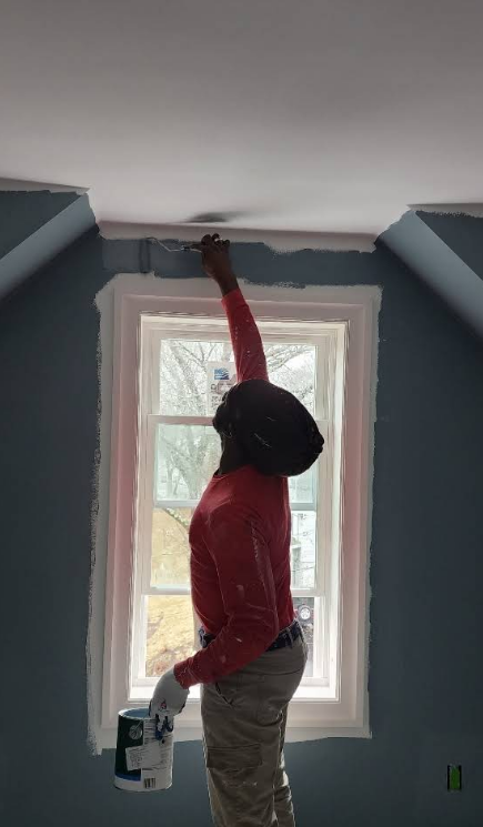 Person painting trim above a window; holding paint can, wearing gloves, in a room with blue walls.