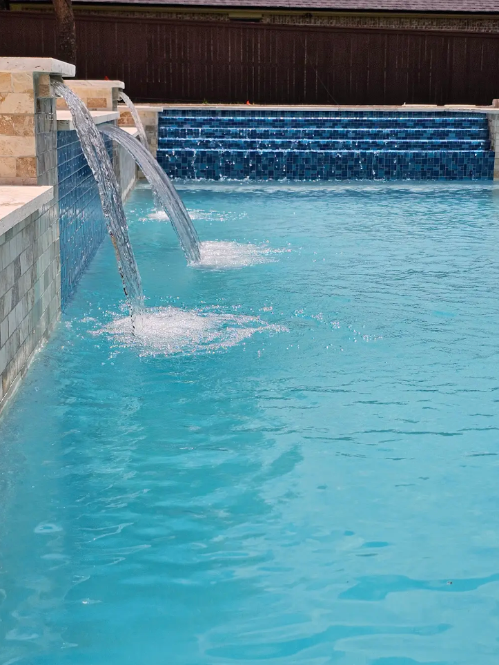 A pool with a raised spa, light blue water, and tan stone coping in a landscaped backyard.