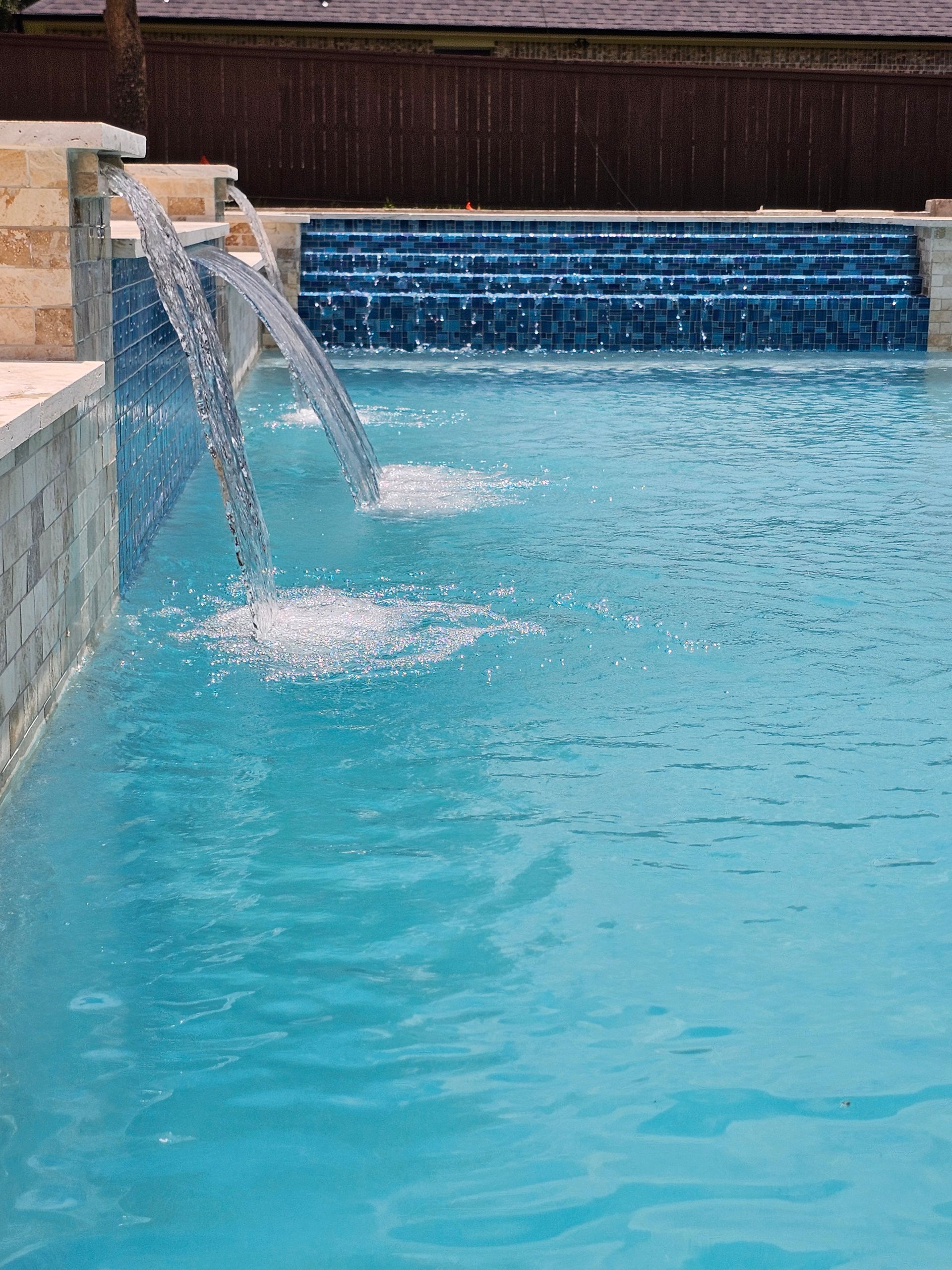 Pool with waterfall feature and built-in stone steps. Turquoise water, rock formation, and outdoor kitchen.