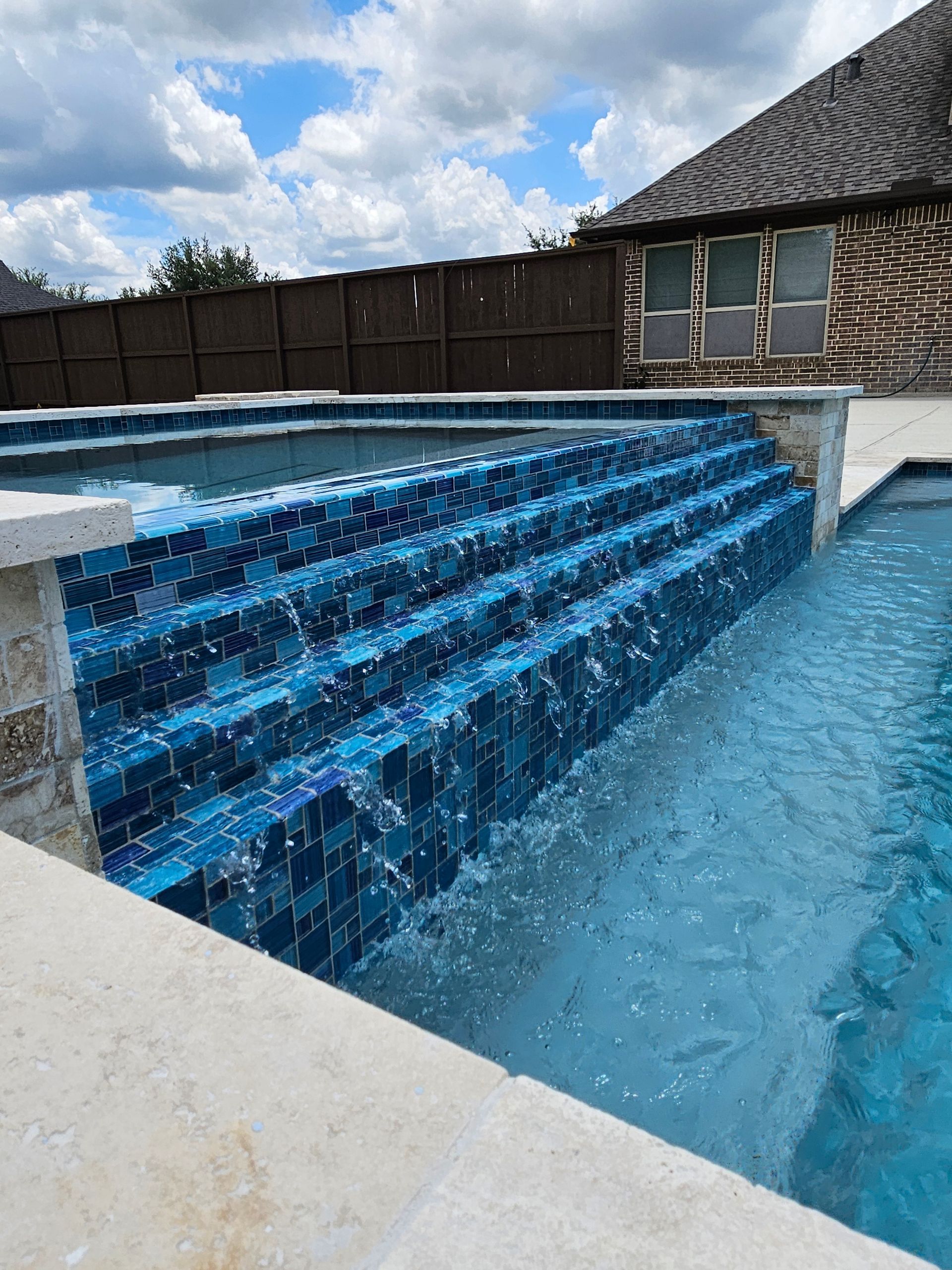 Pool with a spa, water cascading down, patio seating, and green lawn.