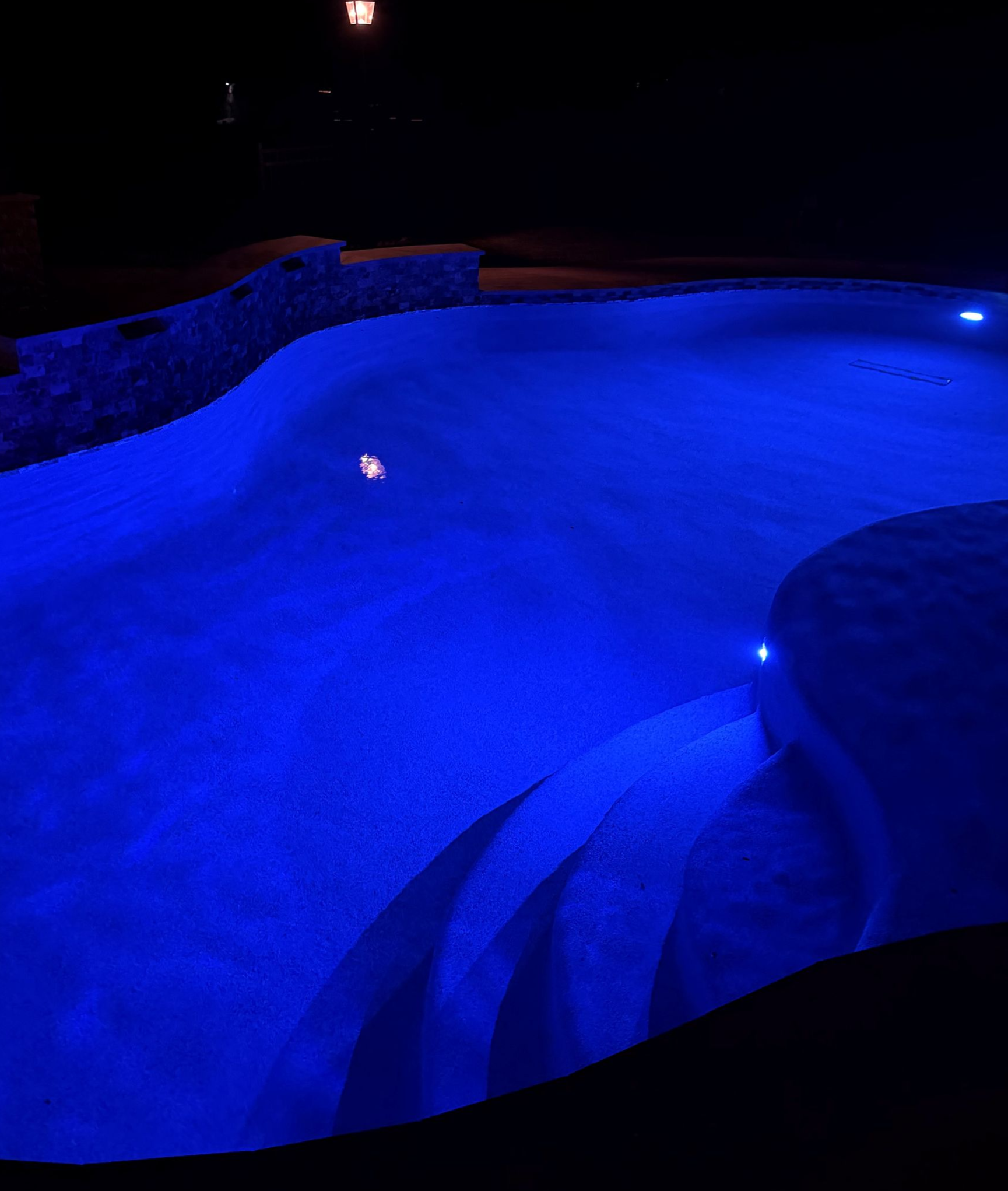 Night view of a pool with blue lights, hot tub, and modern house with outdoor seating.