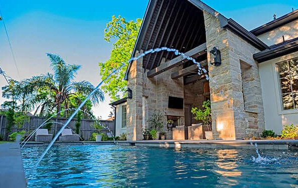 Pool with arching water jets, bordered by green lawn and square stepping stones, outdoor seating area visible.