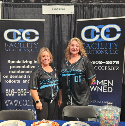 Two women stand at a trade show booth for CC Facility Solutions.