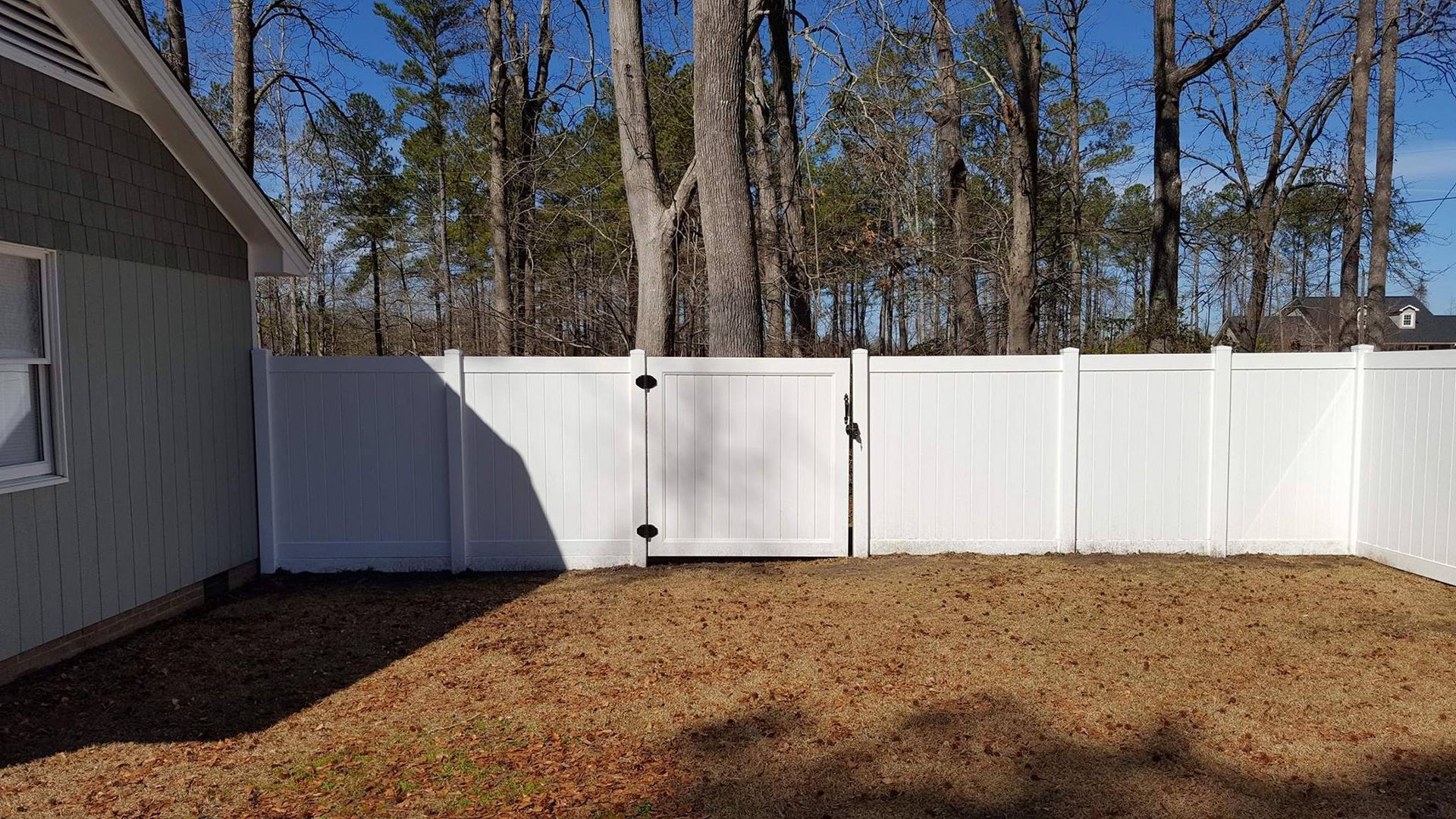 White fenced backyard with a gate, next to a house, brown grass, trees in the background.