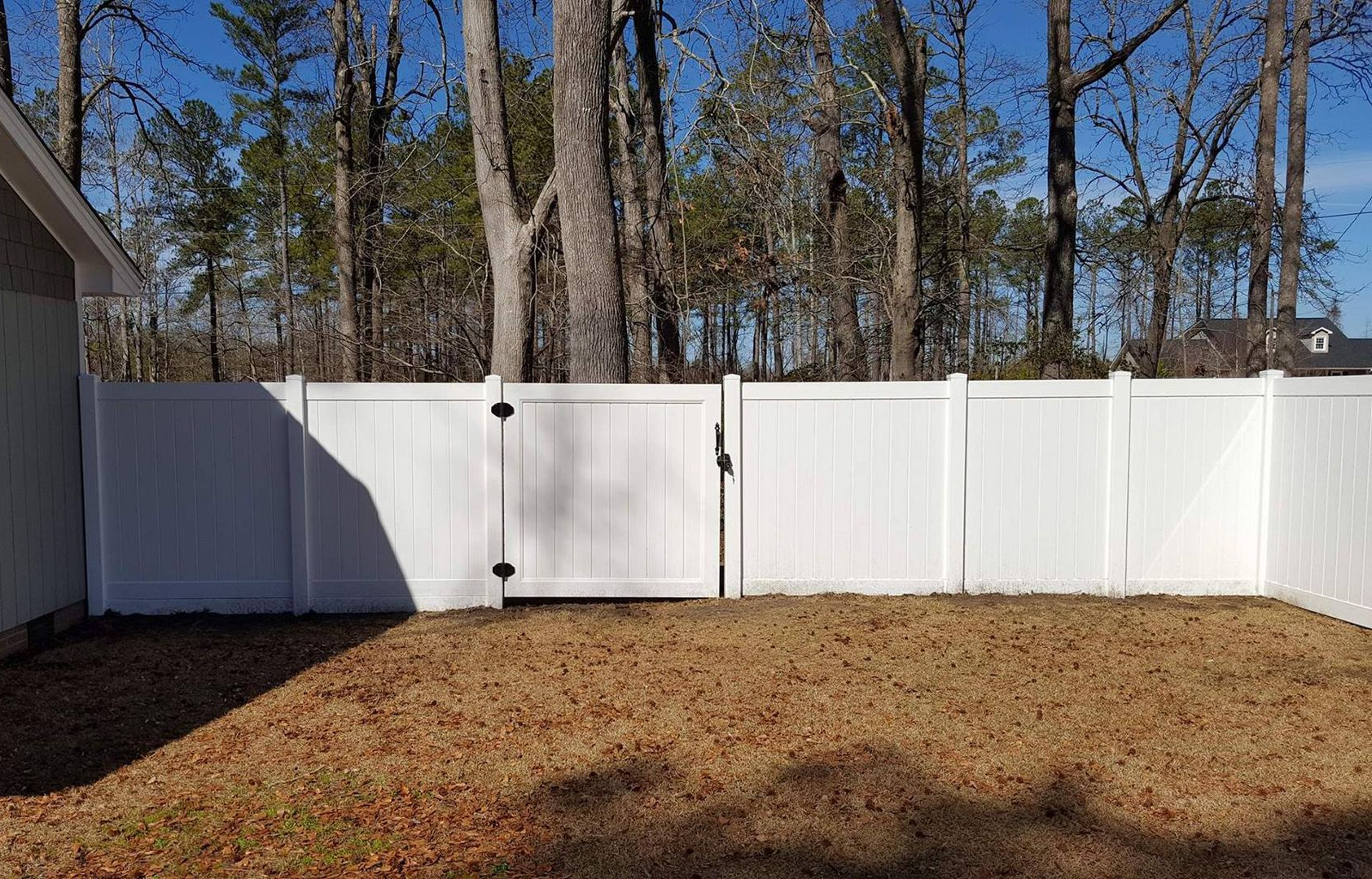 White vinyl fence encloses a backyard with brown grass, gate, and a house on left side. Trees in background.
