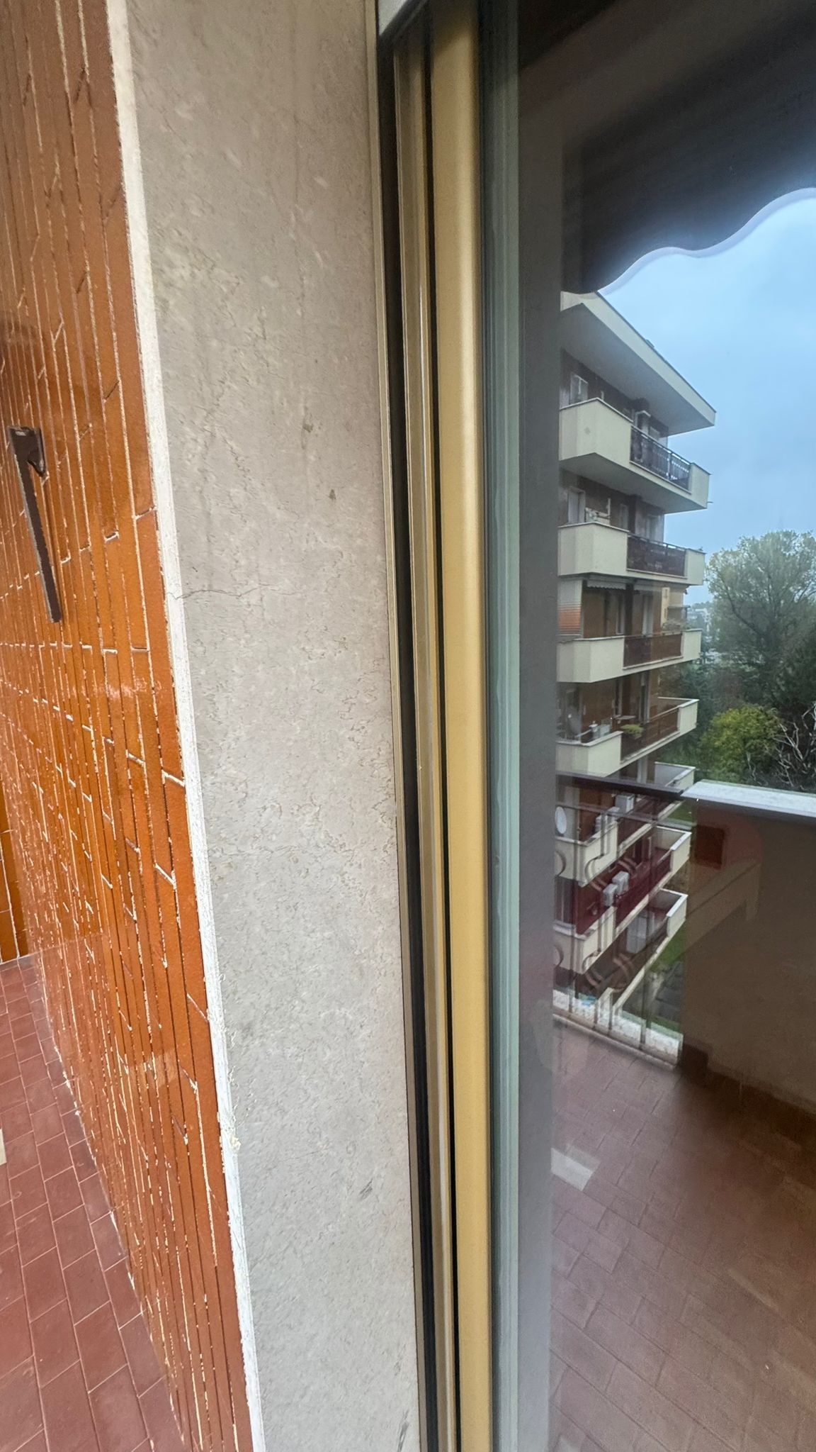Vista dal balcone attraverso la porta scorrevole in vetro; cornice beige, muro di mattoni sulla sinistra.