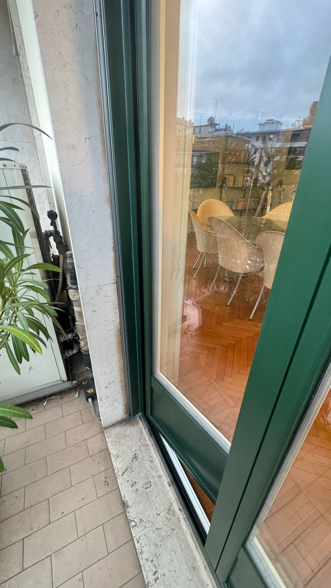 Il telaio verde della finestra, parzialmente aperto, rivela un balcone con sedie e pavimento in legno. 