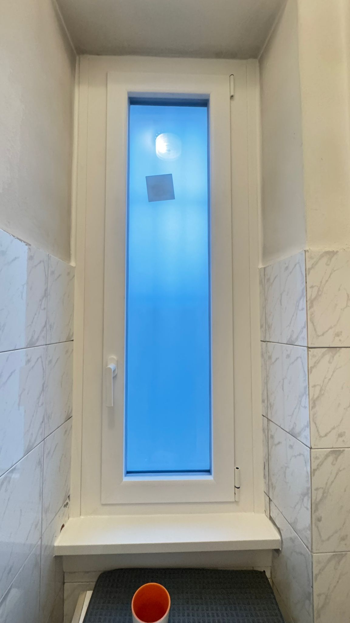 Finestra bianca alta e stretta in un bagno, con vetro smerigliato, piastrelle bianche e un piccolo davanzale.