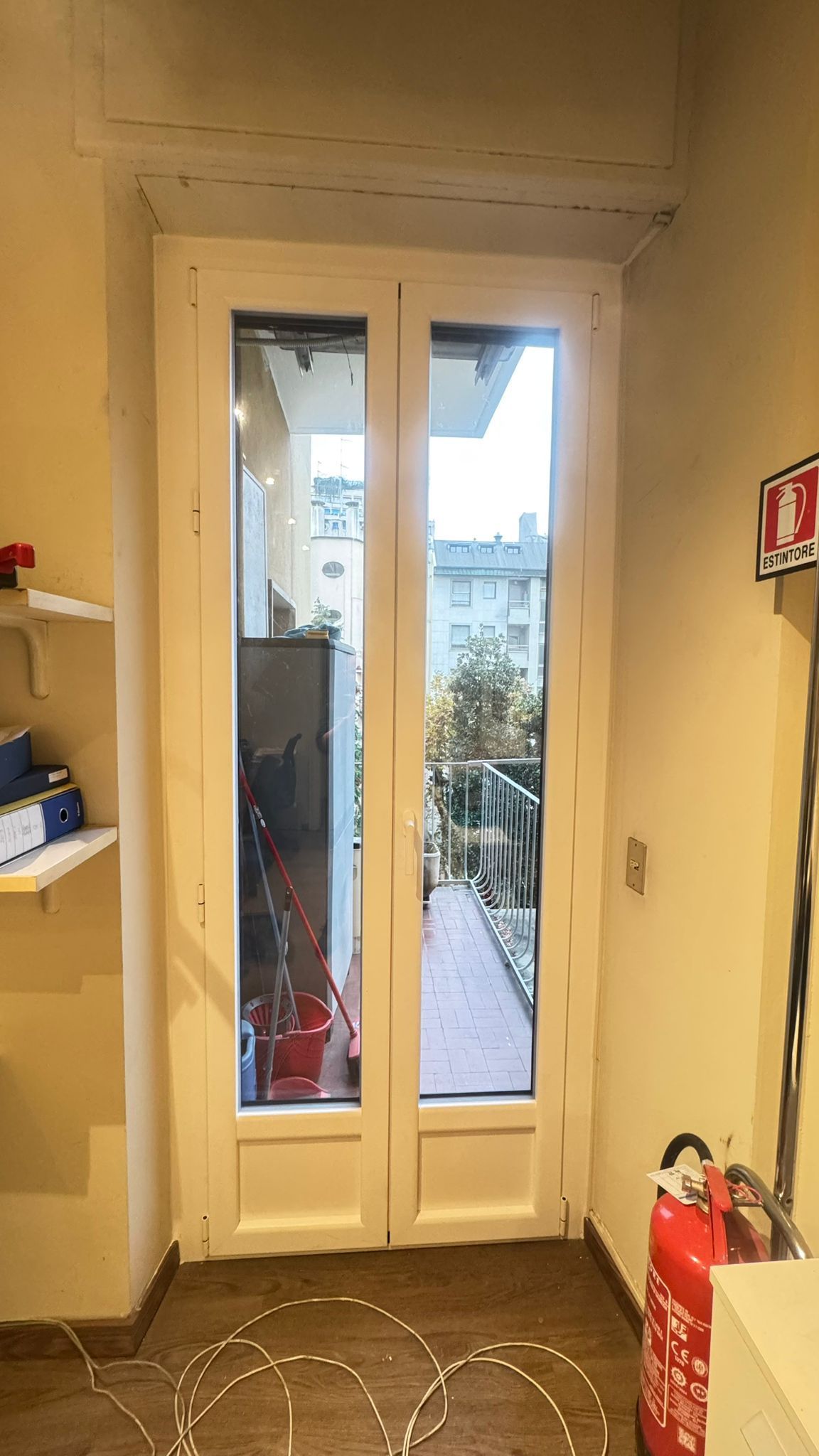Doppie porte a vetri che conducono a un balcone, viste dall'interno. Rifiniture bianche, estintore rosso visibile.