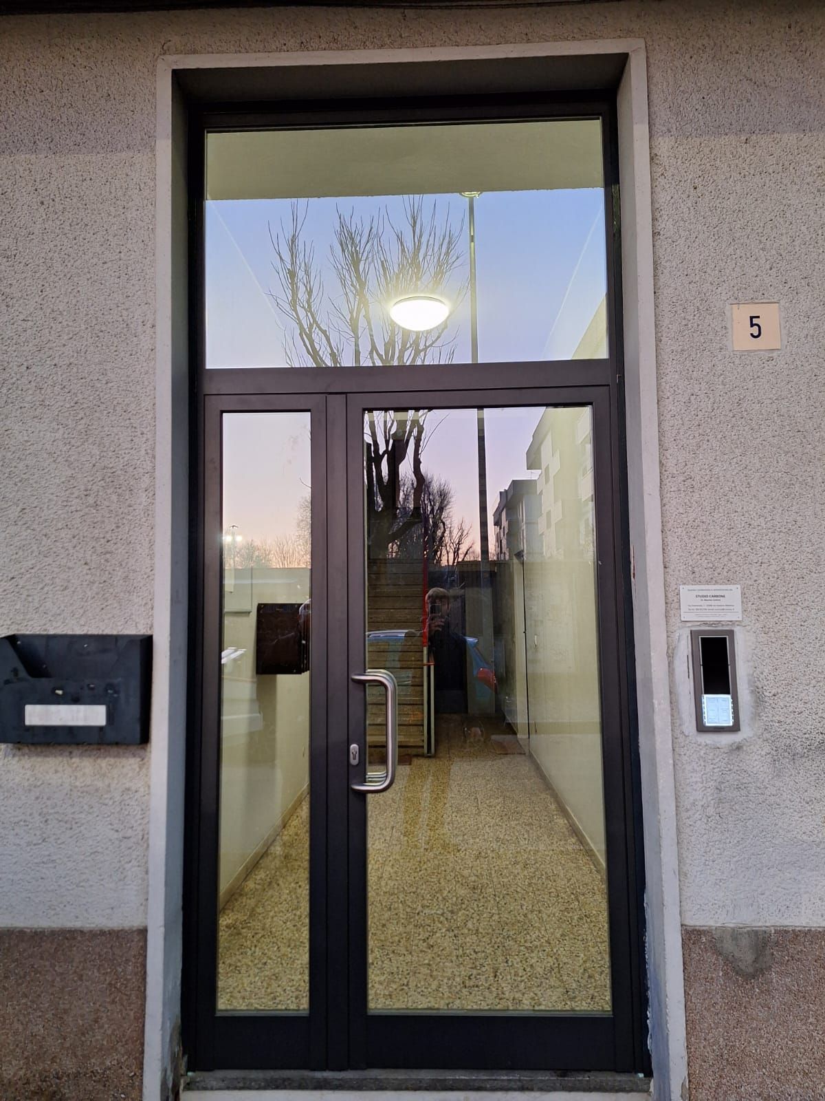 Porta a vetri con vetri laterali, incastonata nella parete esterna grigia. Ingresso visibile dall'interno.