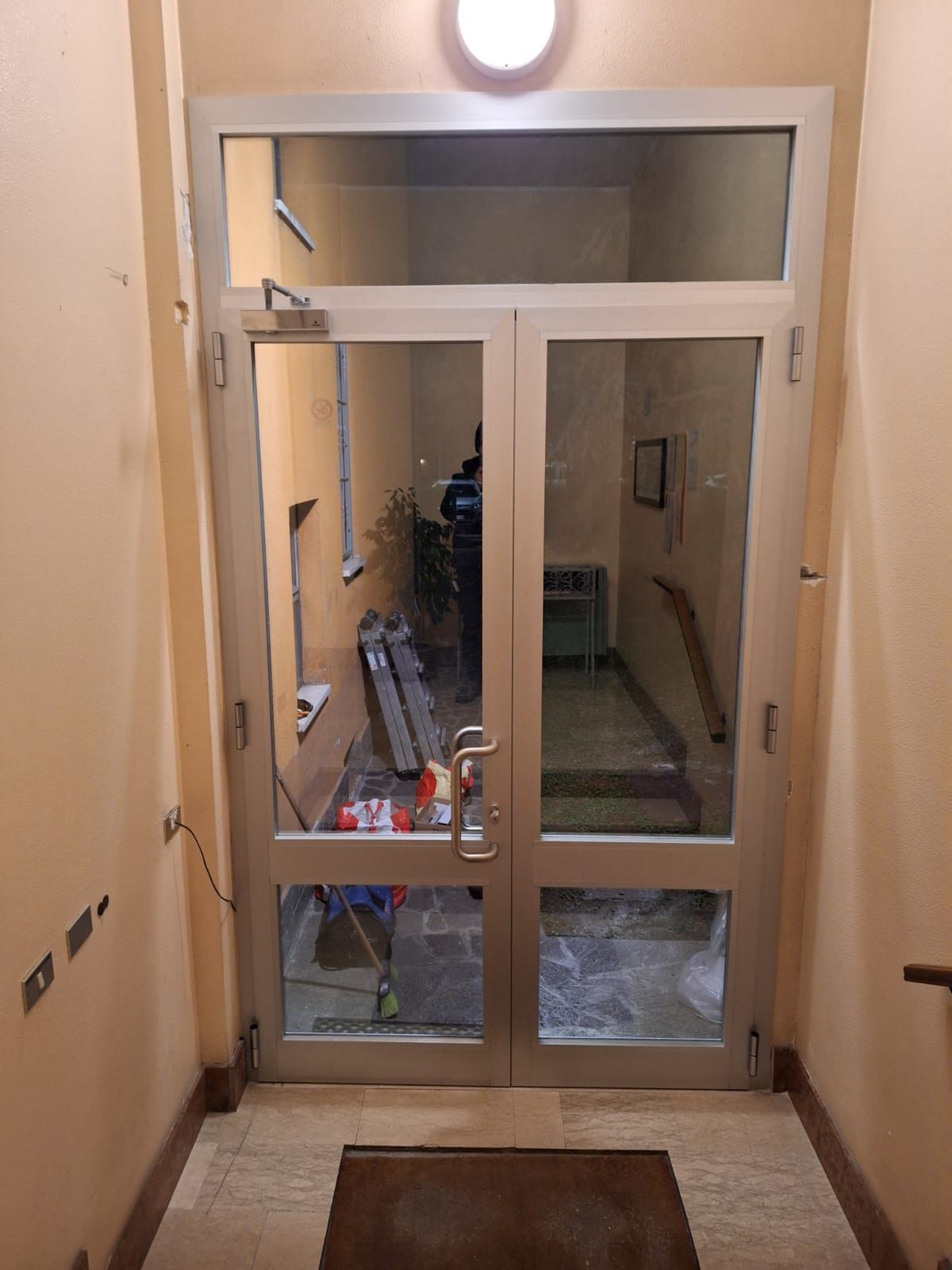 Doppie porte in vetro con telaio in alluminio che conducono a un corridoio con scale e luce zenitale.