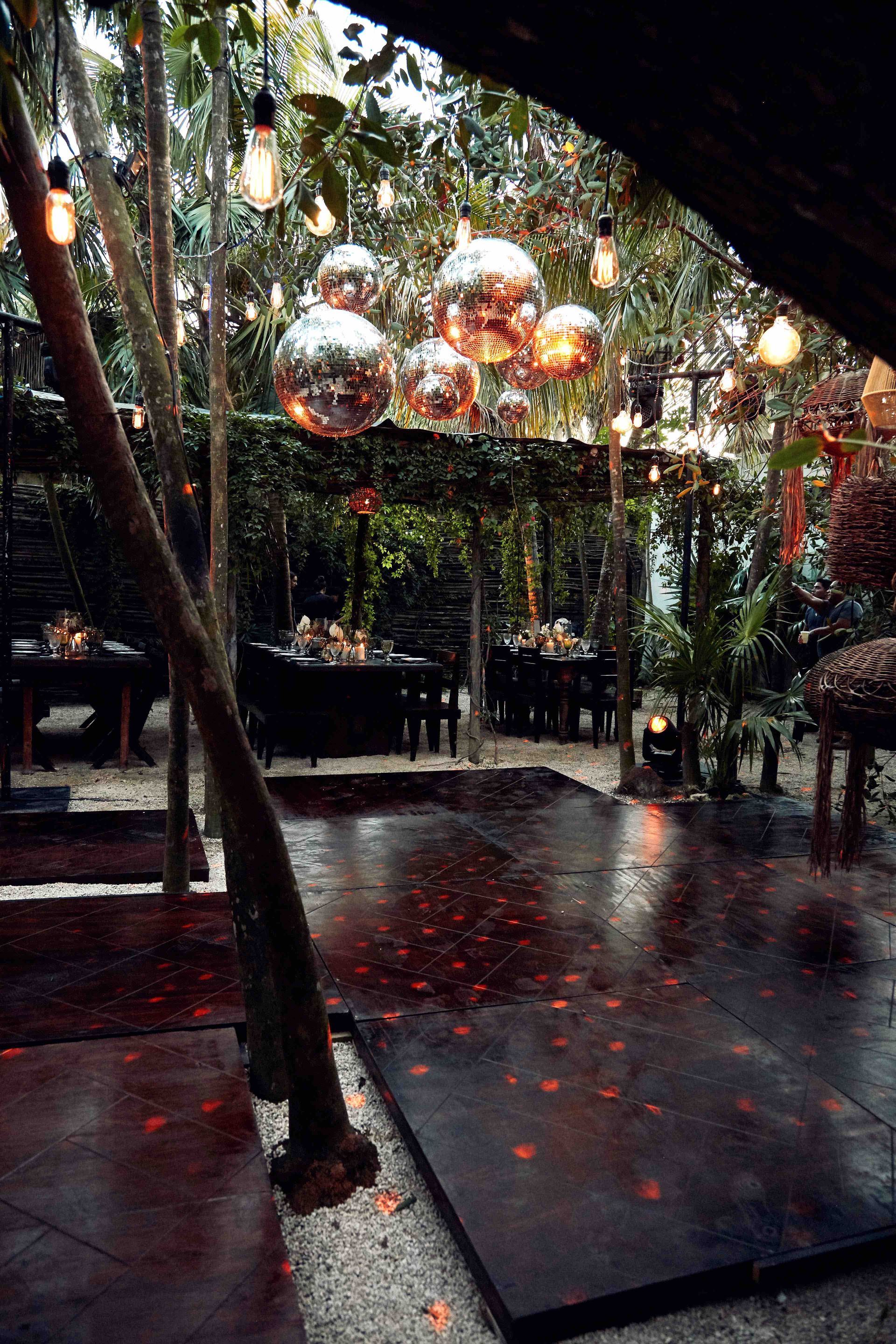Outdoor dining area with disco balls and string lights suspended amongst trees. Red light illuminates the floor.