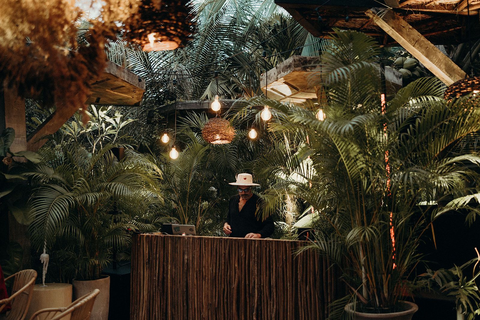 DJ at wooden booth surrounded by tropical plants, string lights overhead.