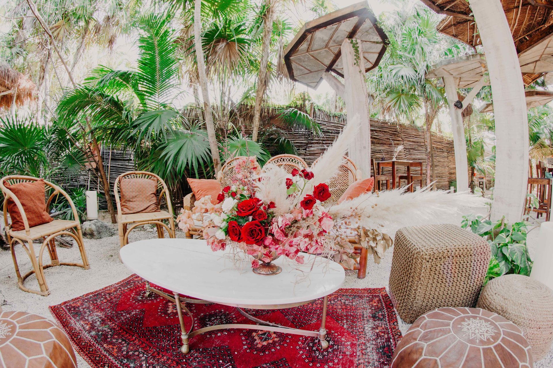 Bohemian outdoor lounge area with wicker furniture, red rug, and flower arrangement.