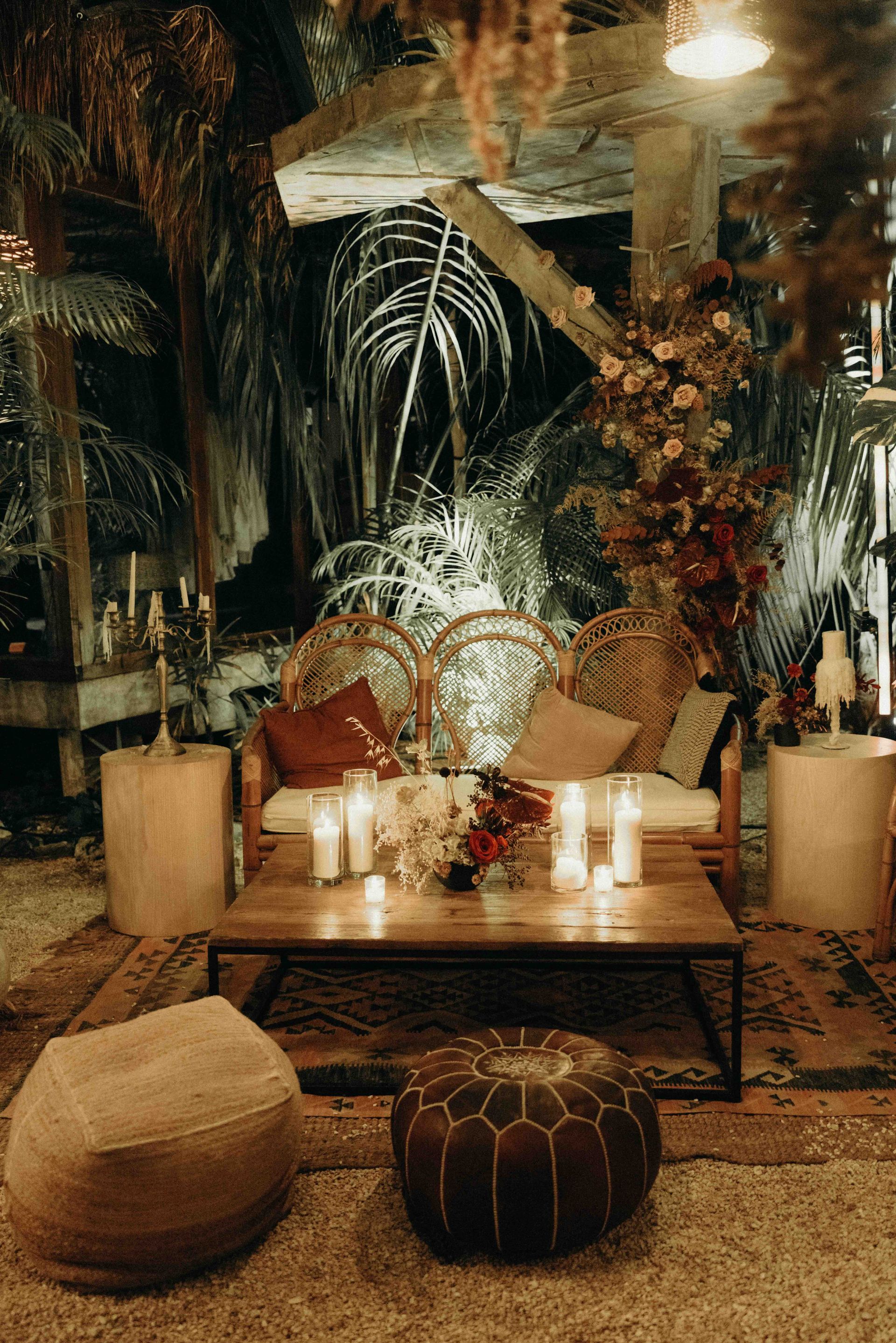 Cozy outdoor seating area with candles, pillows, and rugs. Wooden table, poufs, and lush greenery.