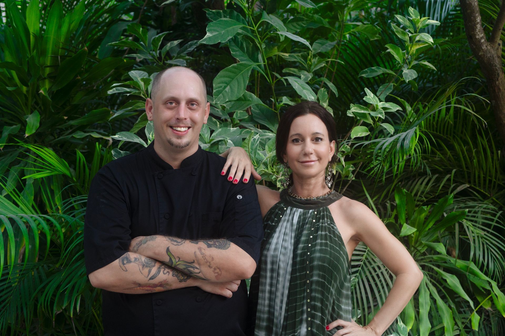 A man and a woman are posing for a picture in front of a lush green forest.