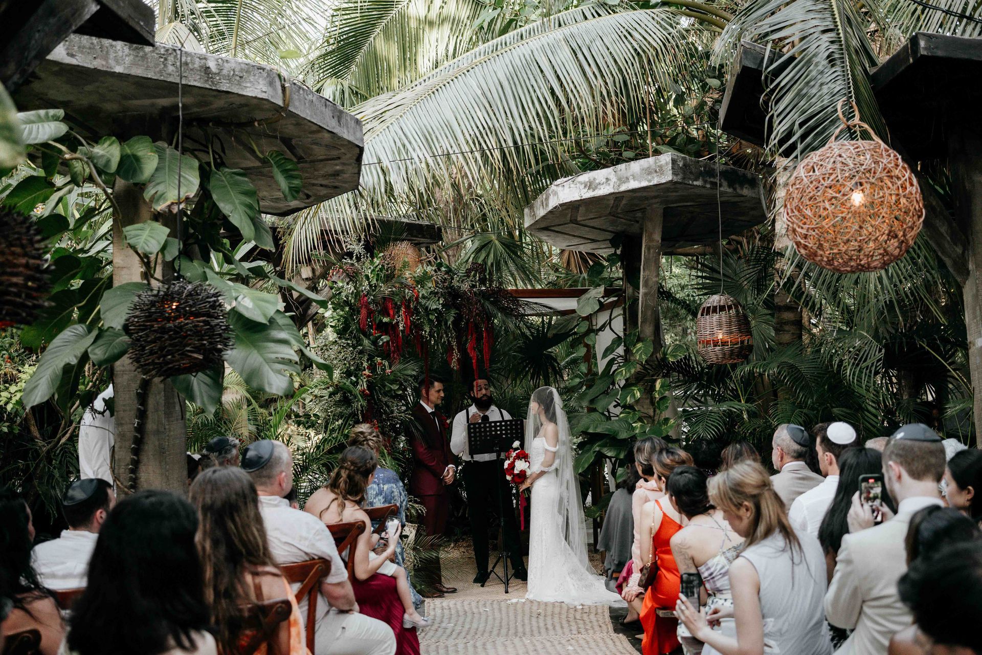 Wedding ceremony in lush tropical garden, bride & groom at altar, guests seated.