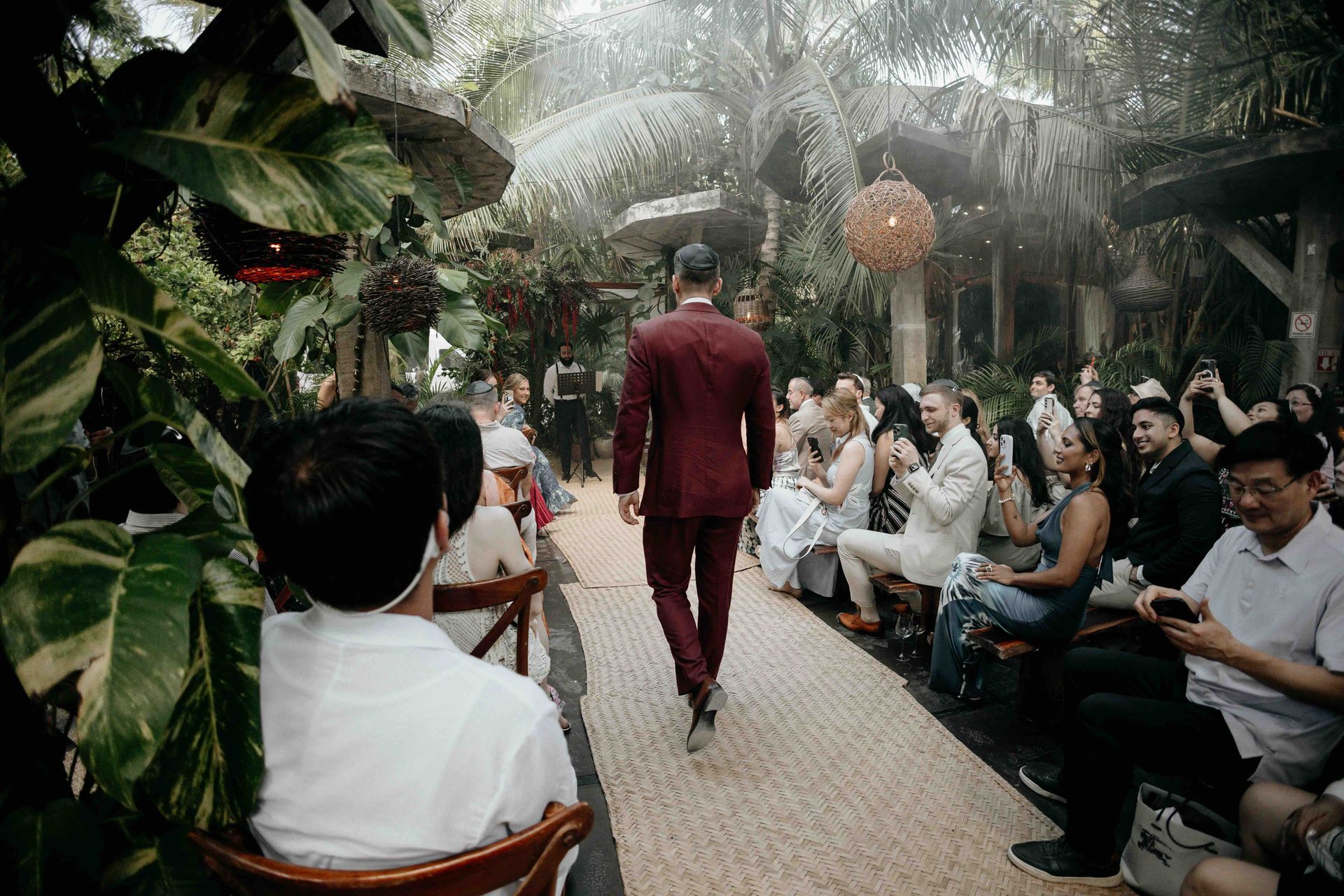 Man in burgundy suit walks down aisle towards wedding guests in lush, outdoor setting.