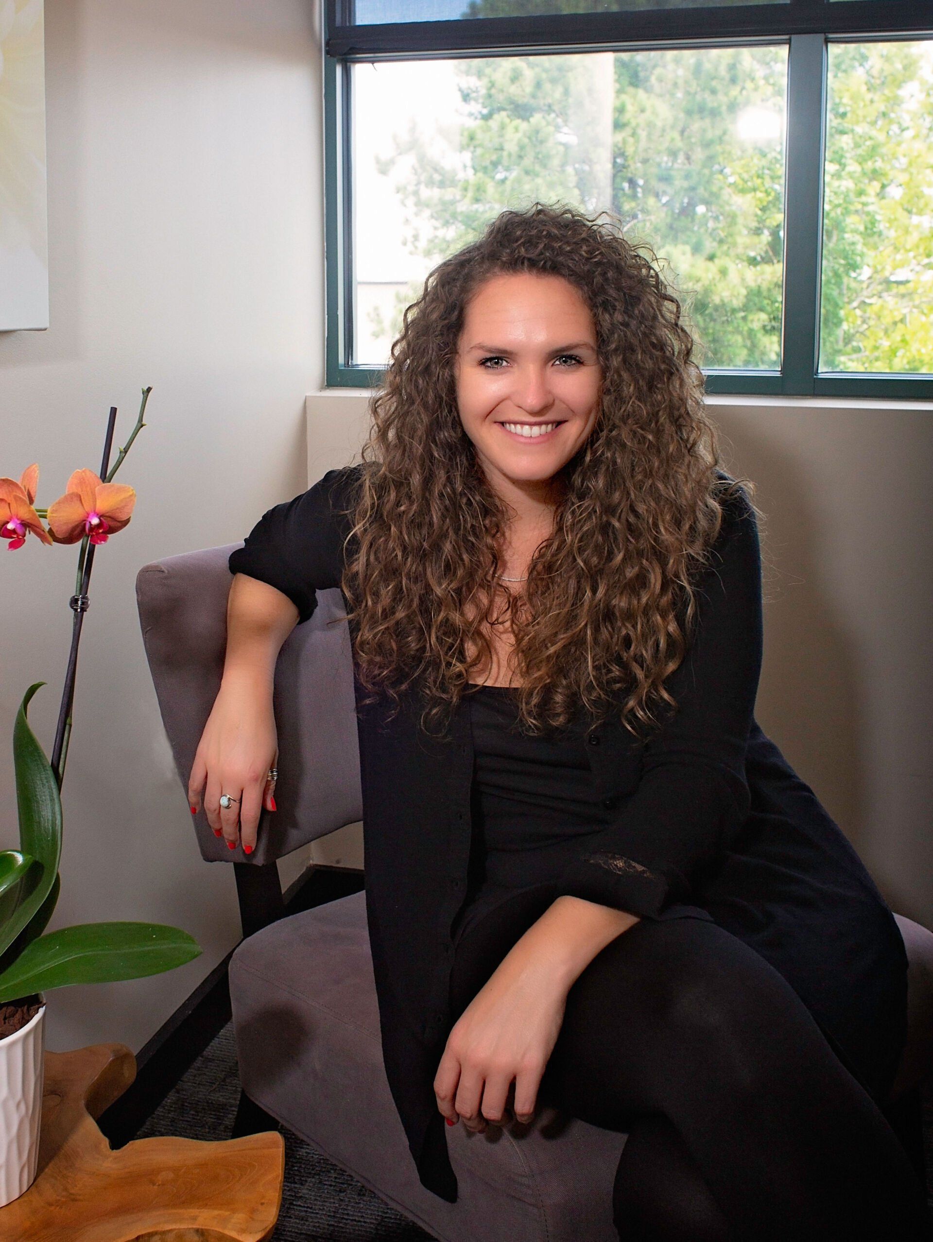 A woman with curly hair is sitting in a chair in front of a window.