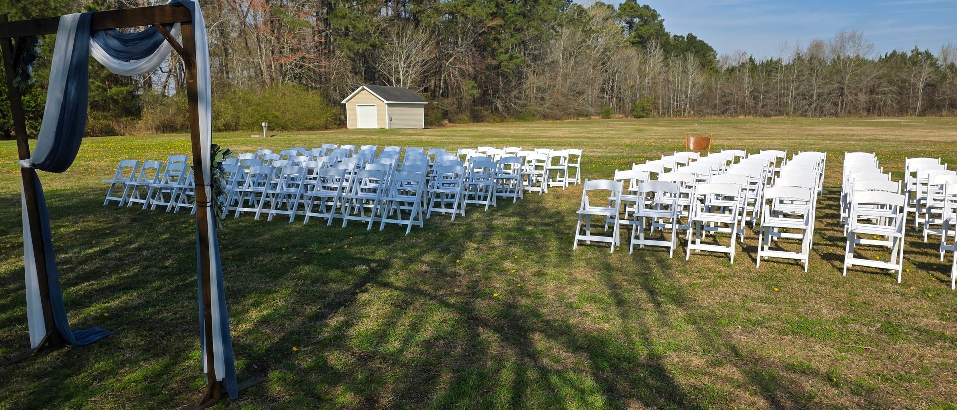 Rows of white folding chairs set up for an outdoor wedding ceremony in a grassy field with a wooden arch in the foreground.