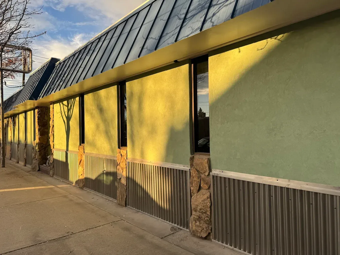 Sunlit storefront with yellow panels, corrugated metal base, and a glass awning along a sidewalk