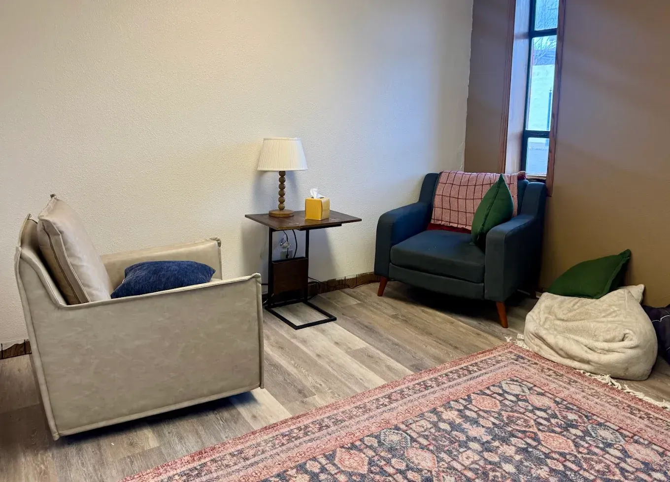Small cozy living room with beige and green chairs, a lamp, side table, rug, and floor pillows by a window