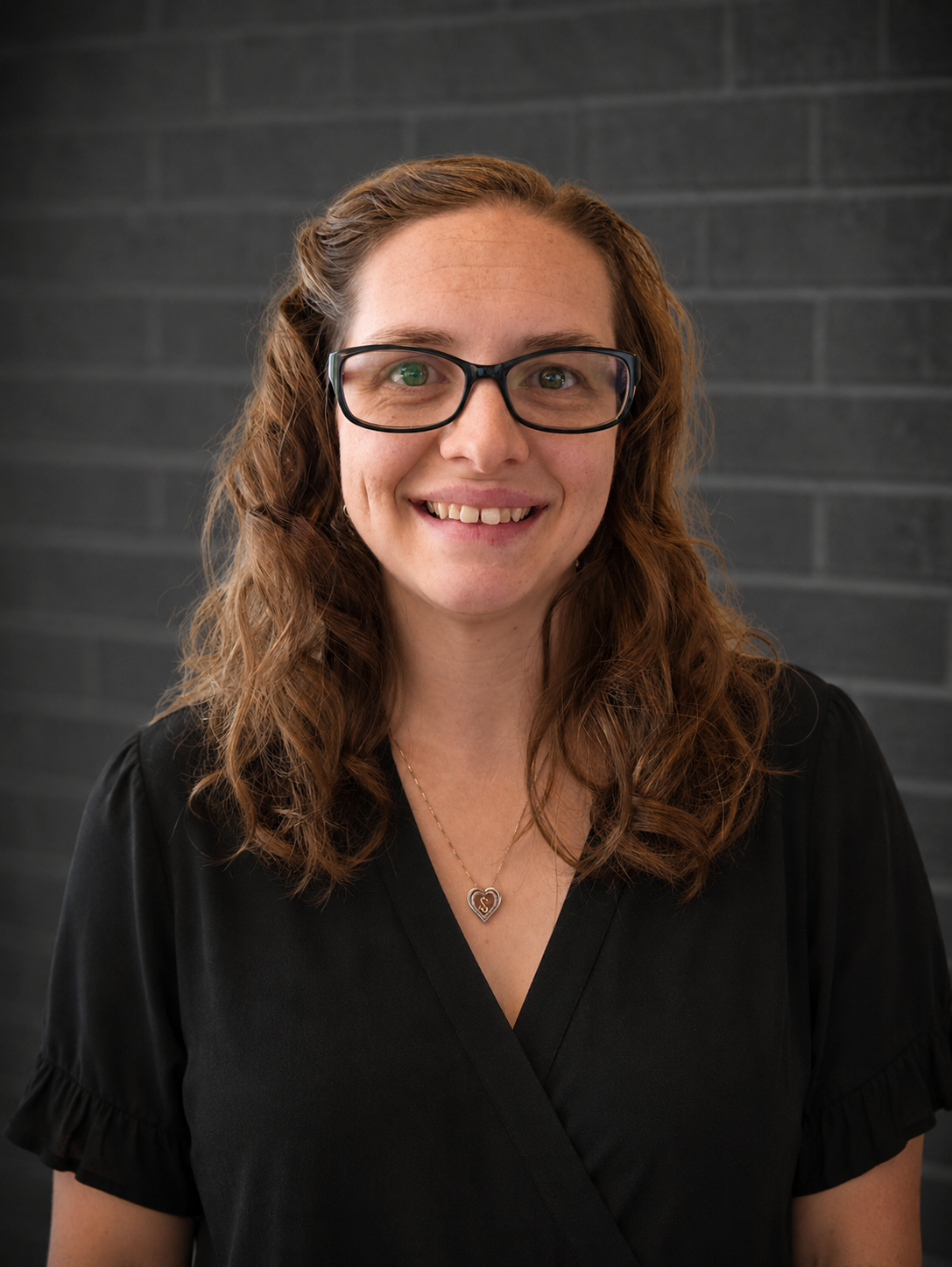 Portrait of a smiling person with glasses and curly hair, wearing a black blouse against a dark brick wall.