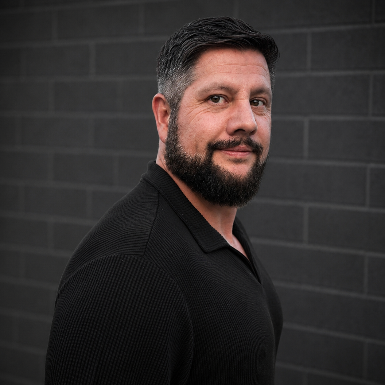 Portrait of a bearded man in a black shirt against a dark brick wall