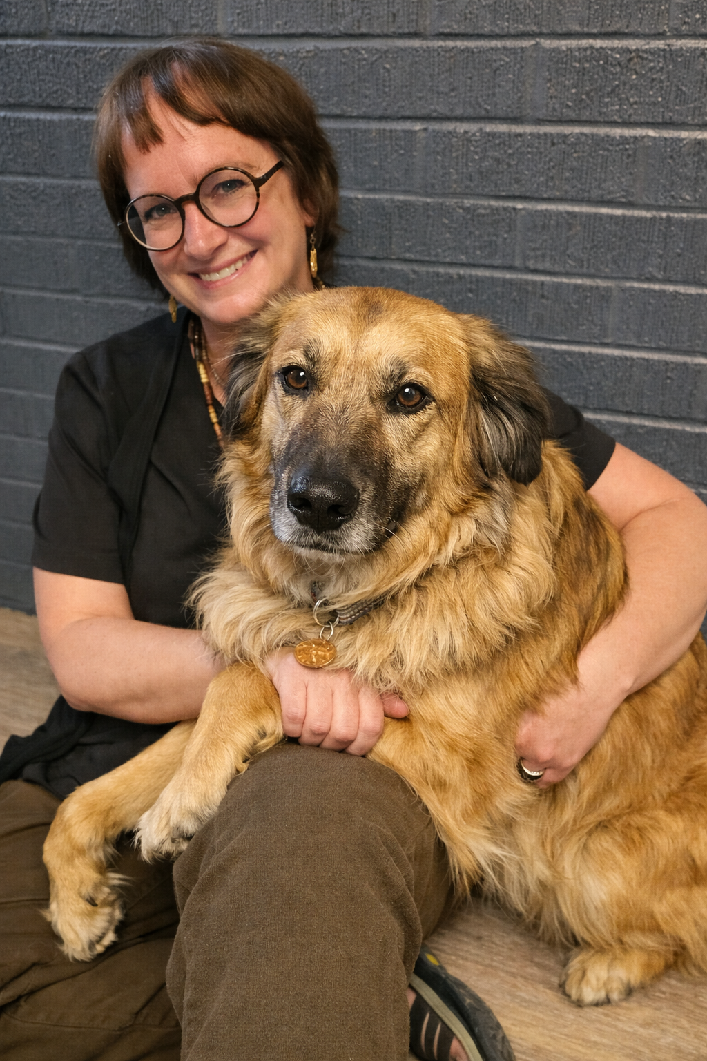 Woman in glasses hugging a large tan dog against a gray wall