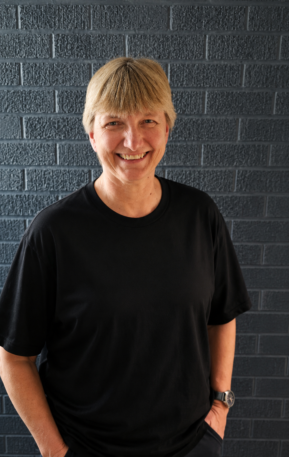 Smiling person in a black shirt standing against a dark brick wall