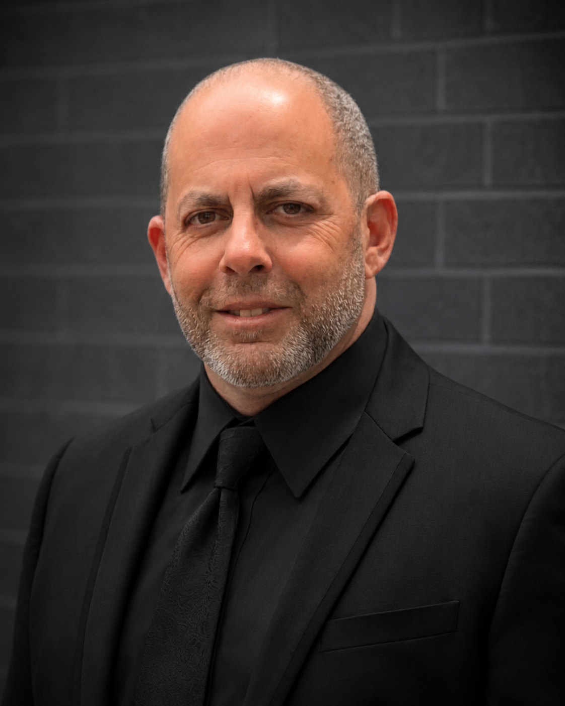 Portrait of a bald man in a black suit and tie, standing against a dark brick wall with a slight smile