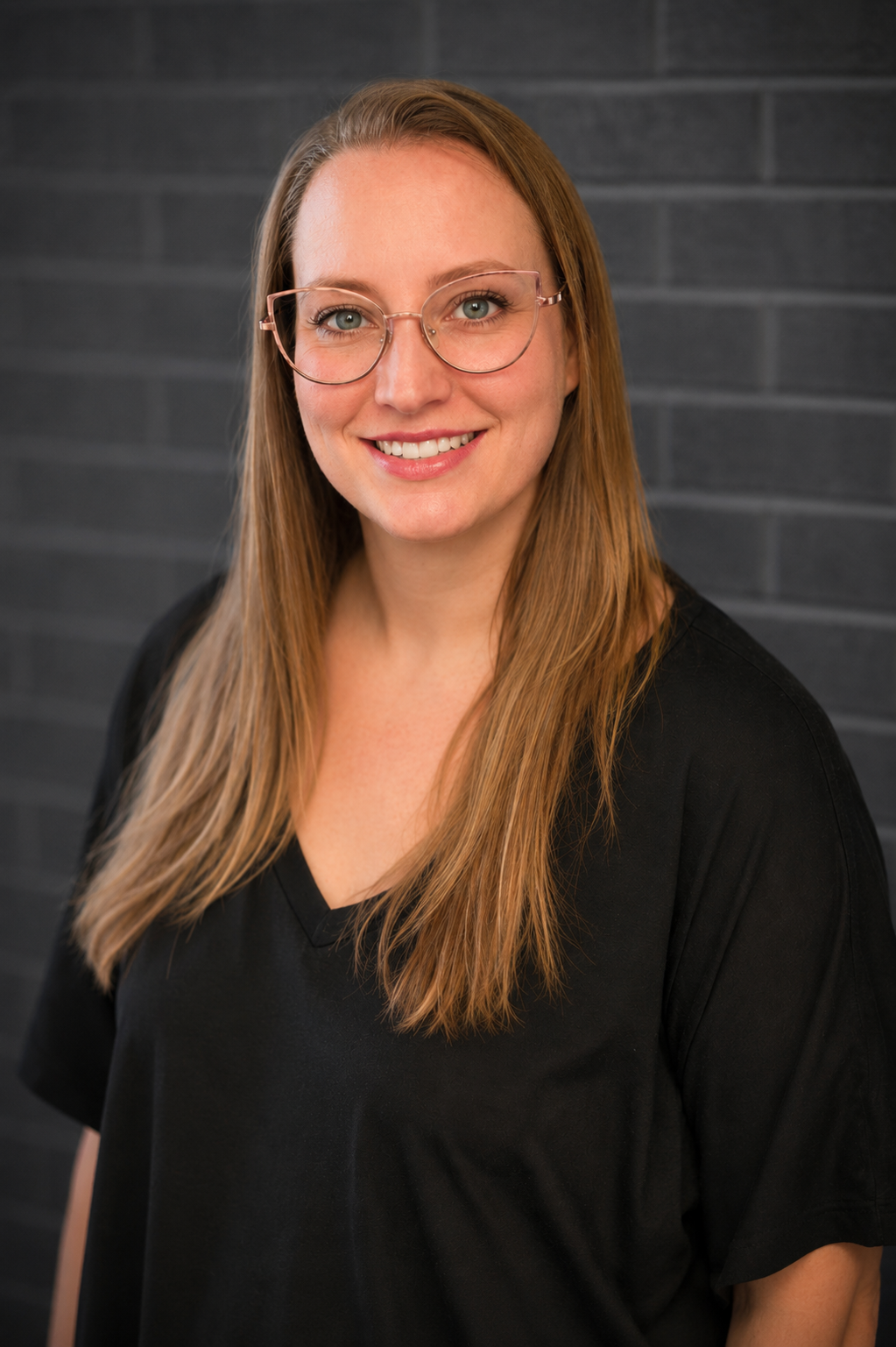 Smiling woman with glasses and long hair in a black top against a dark brick wall