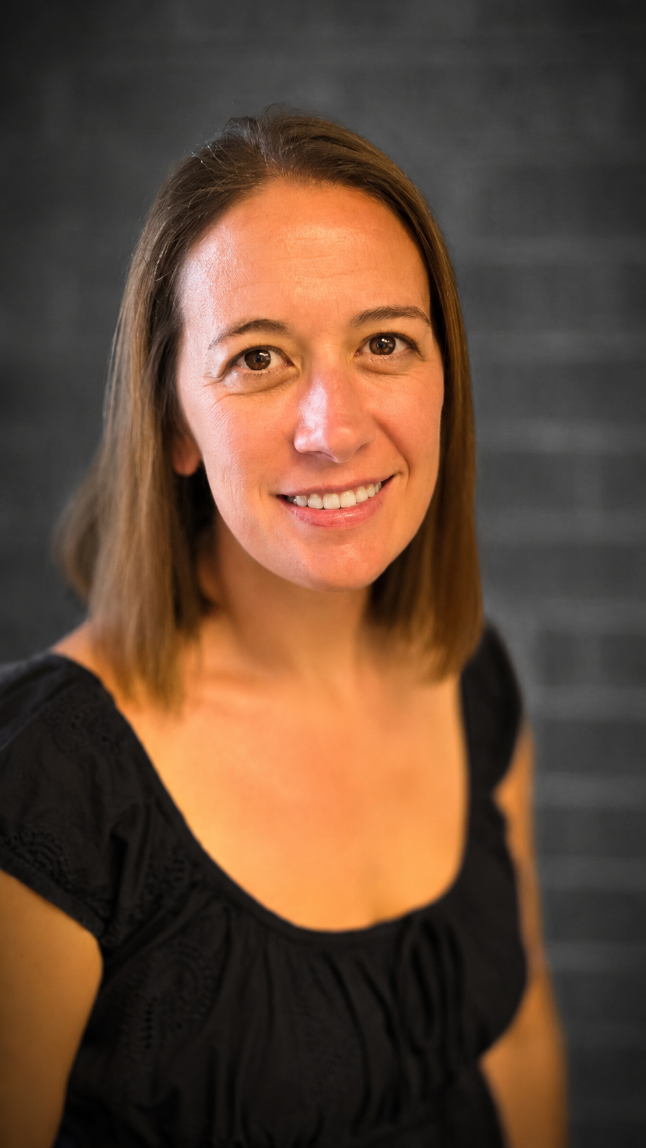 Portrait of a smiling person with shoulder-length brown hair against a dark gray backdrop