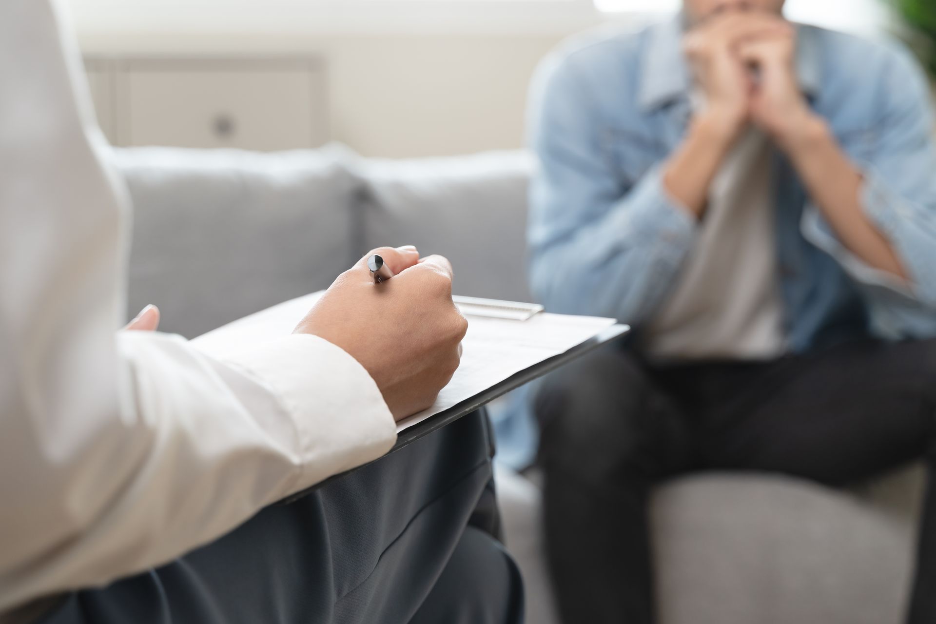 Therapist taking notes while a client sits on a couch, hands clasped in a 상담 room.