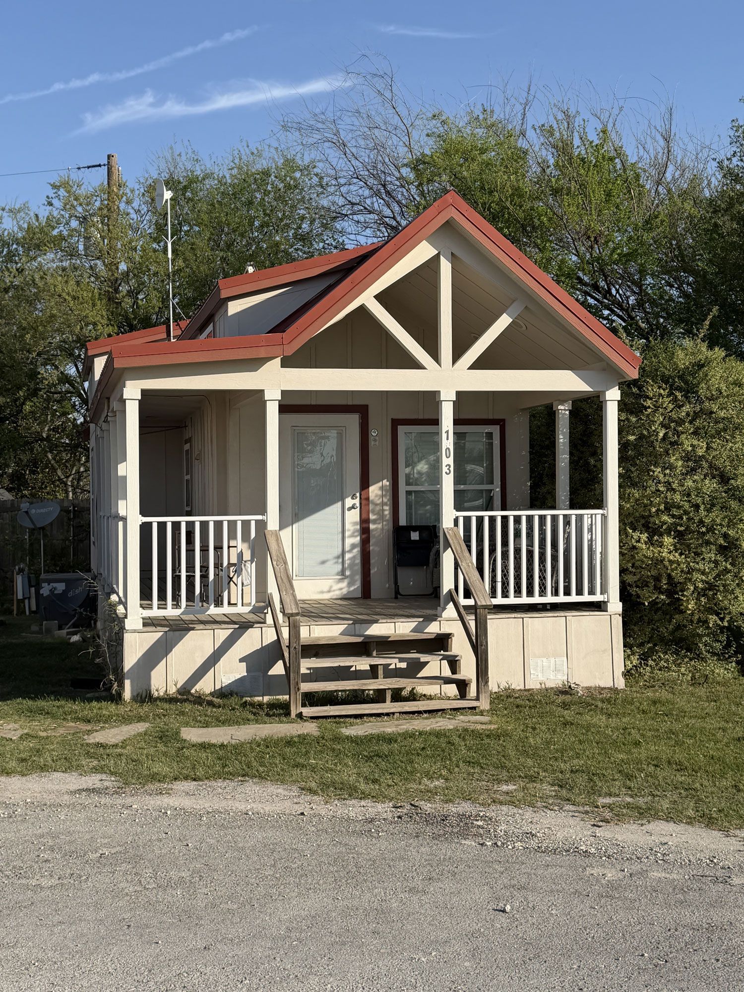 Small beige cottage with red roof, front porch, and steps beside a gravel road.