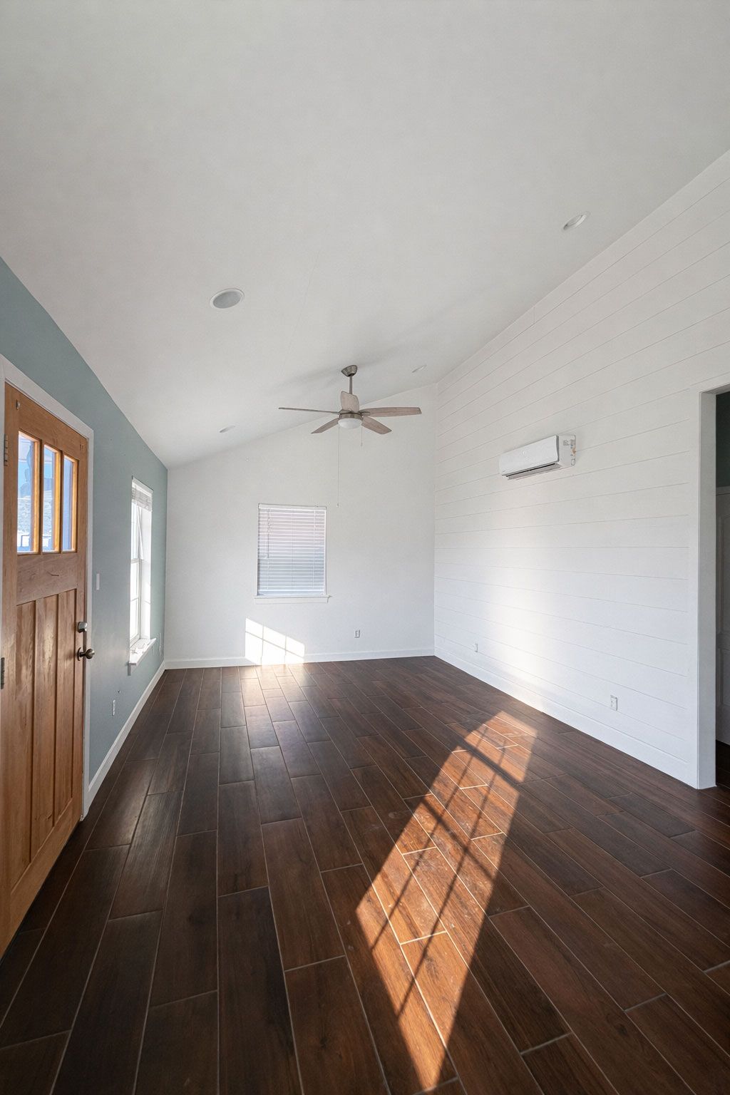 Empty room with dark hardwood floors, white walls, ceiling fan, and sunlight casting long shadows.