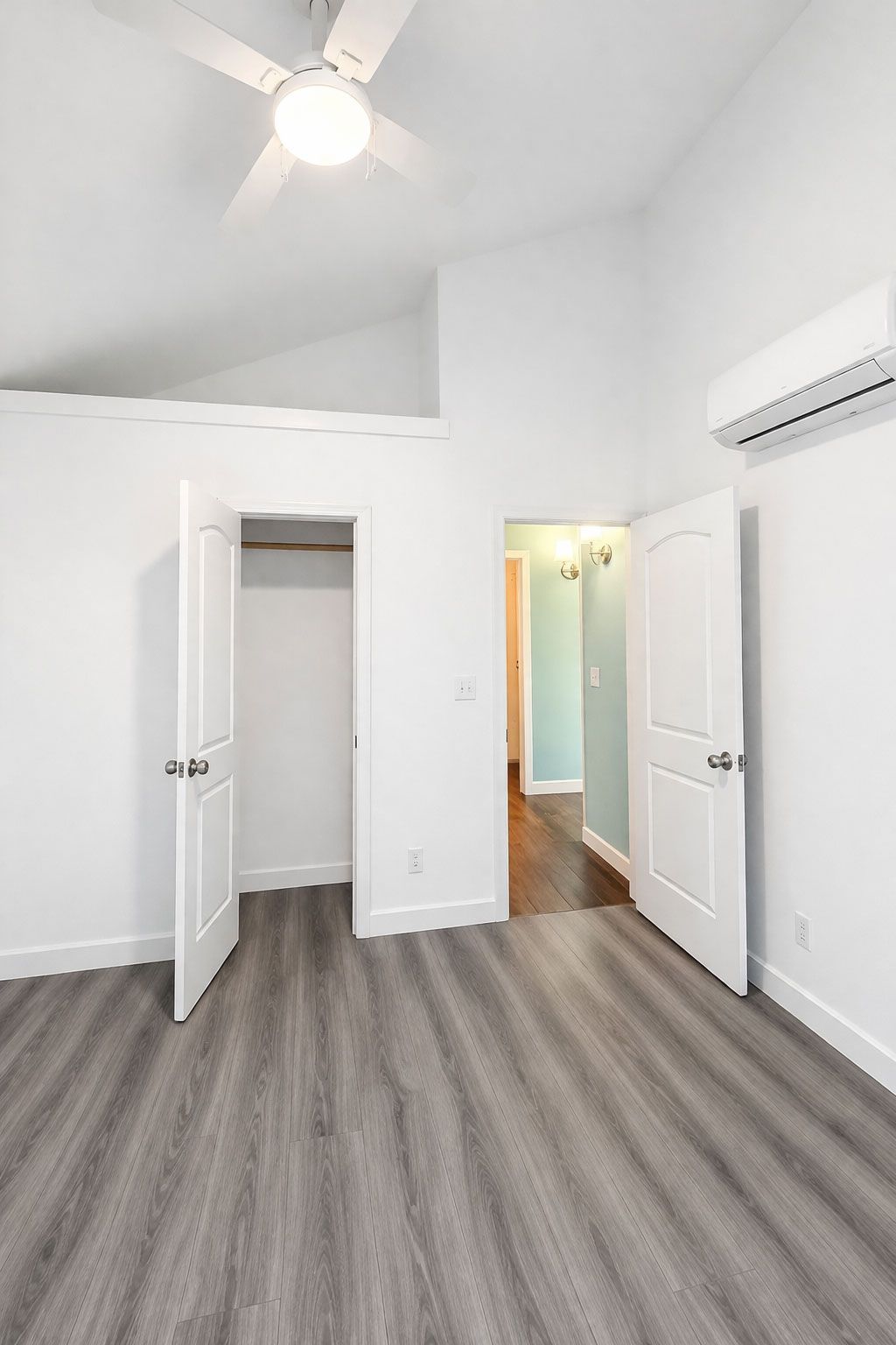 Bright empty bedroom with gray carpet, white walls, open doors, and a ceiling fan.