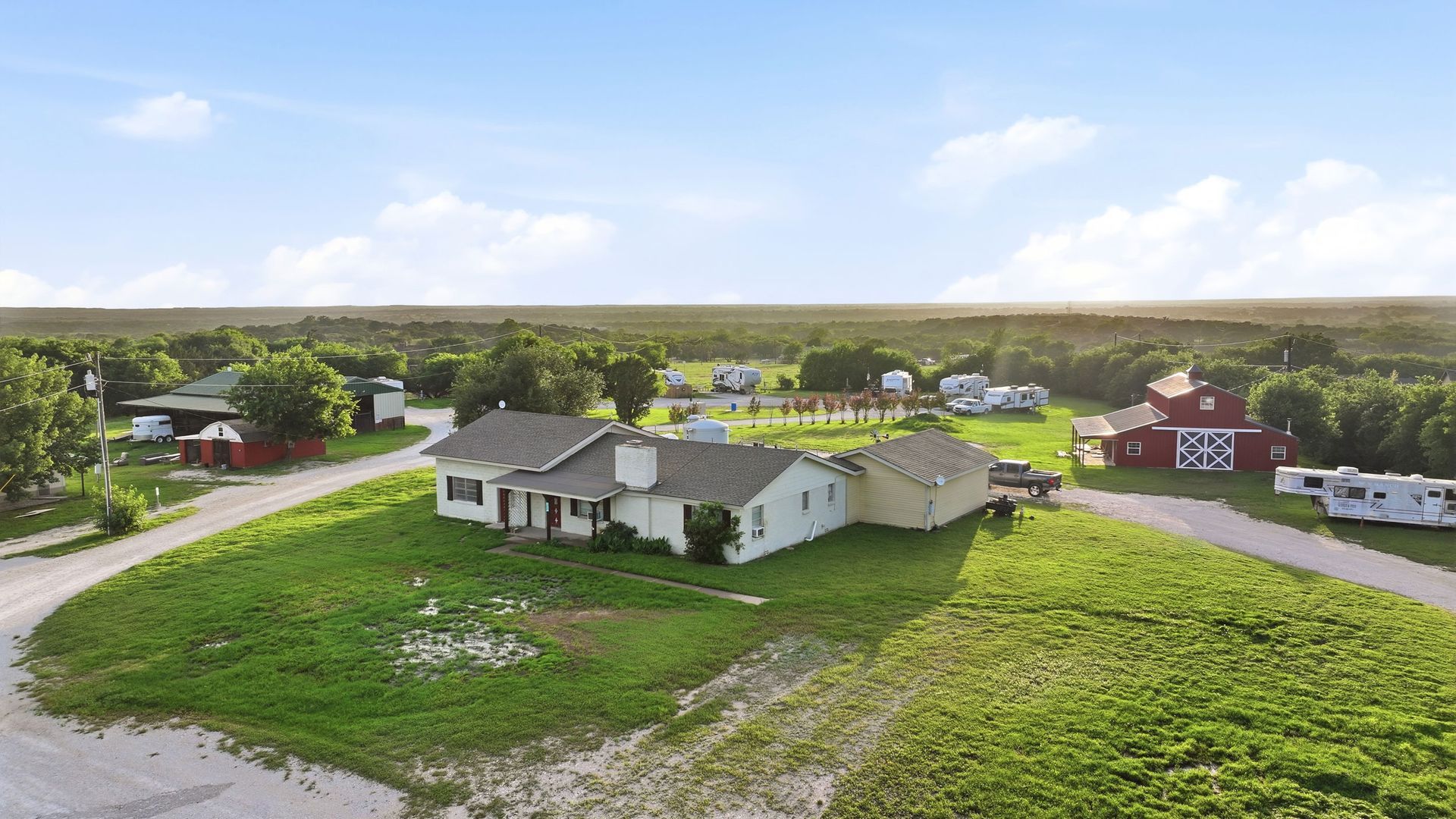 Aerial view of a white farmhouse and outbuildings on a green rural property under a bright sky