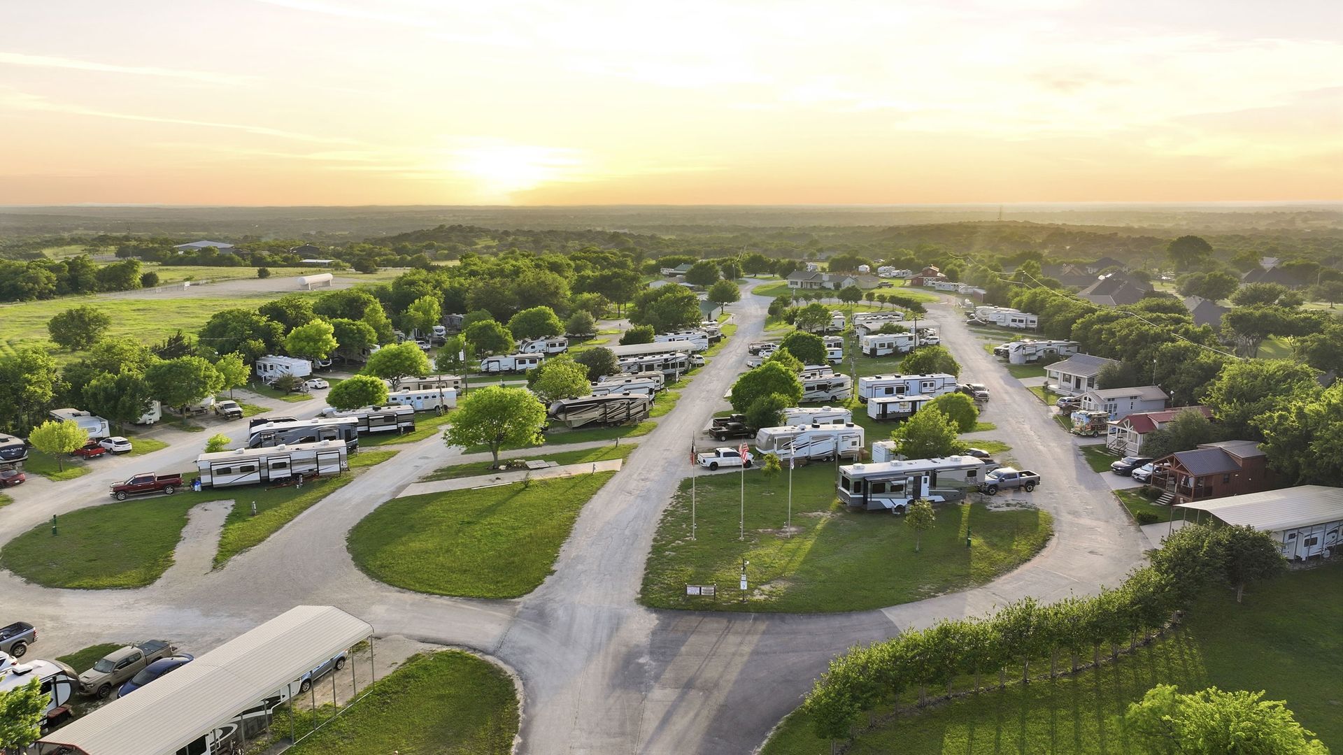 Aerial view of a suburban RV park at sunset, with rows of trailers, roads, and green trees.